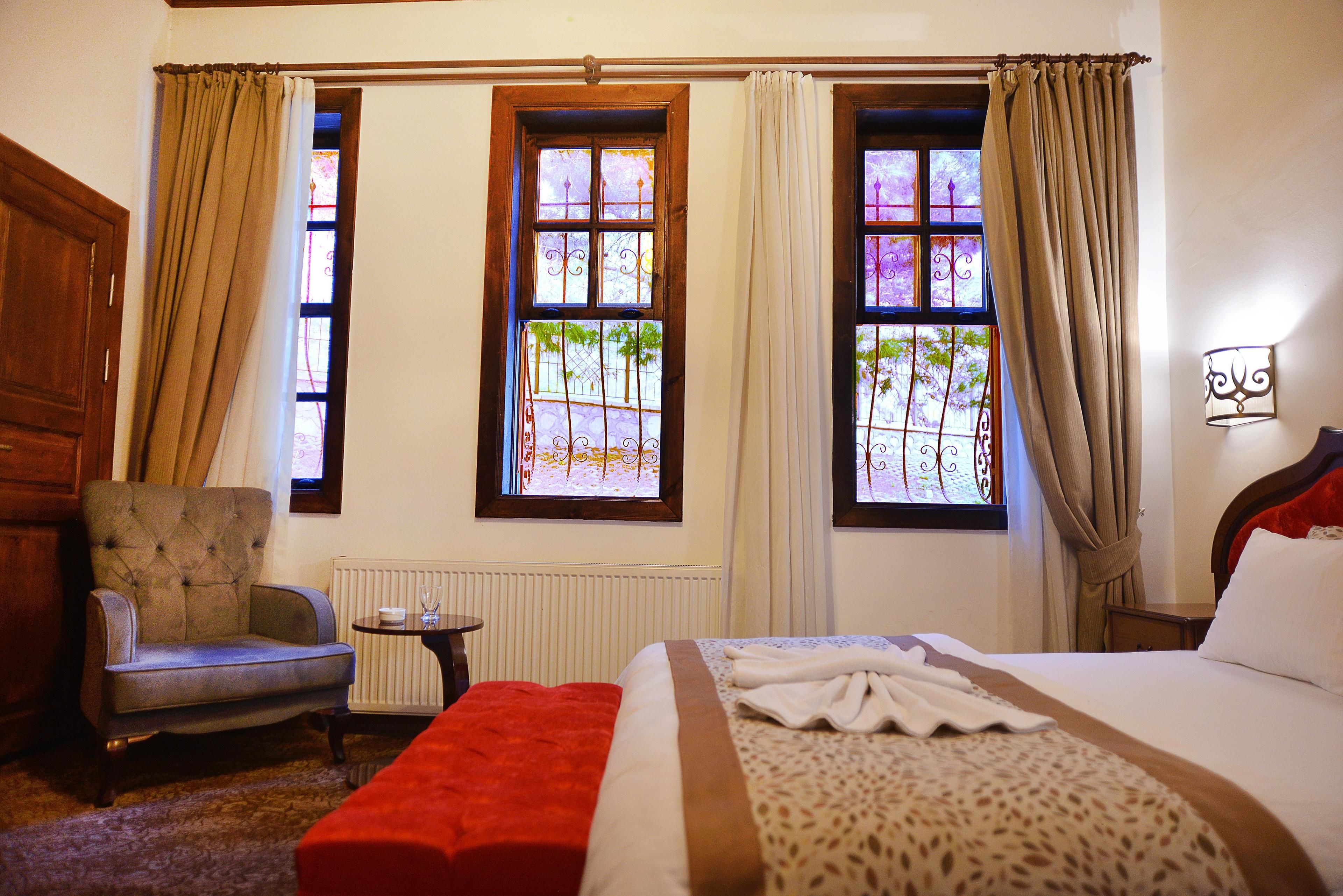 Quarto de hotel com uma cama, uma cadeira, uma mesinha e duas janelas com cortinas.