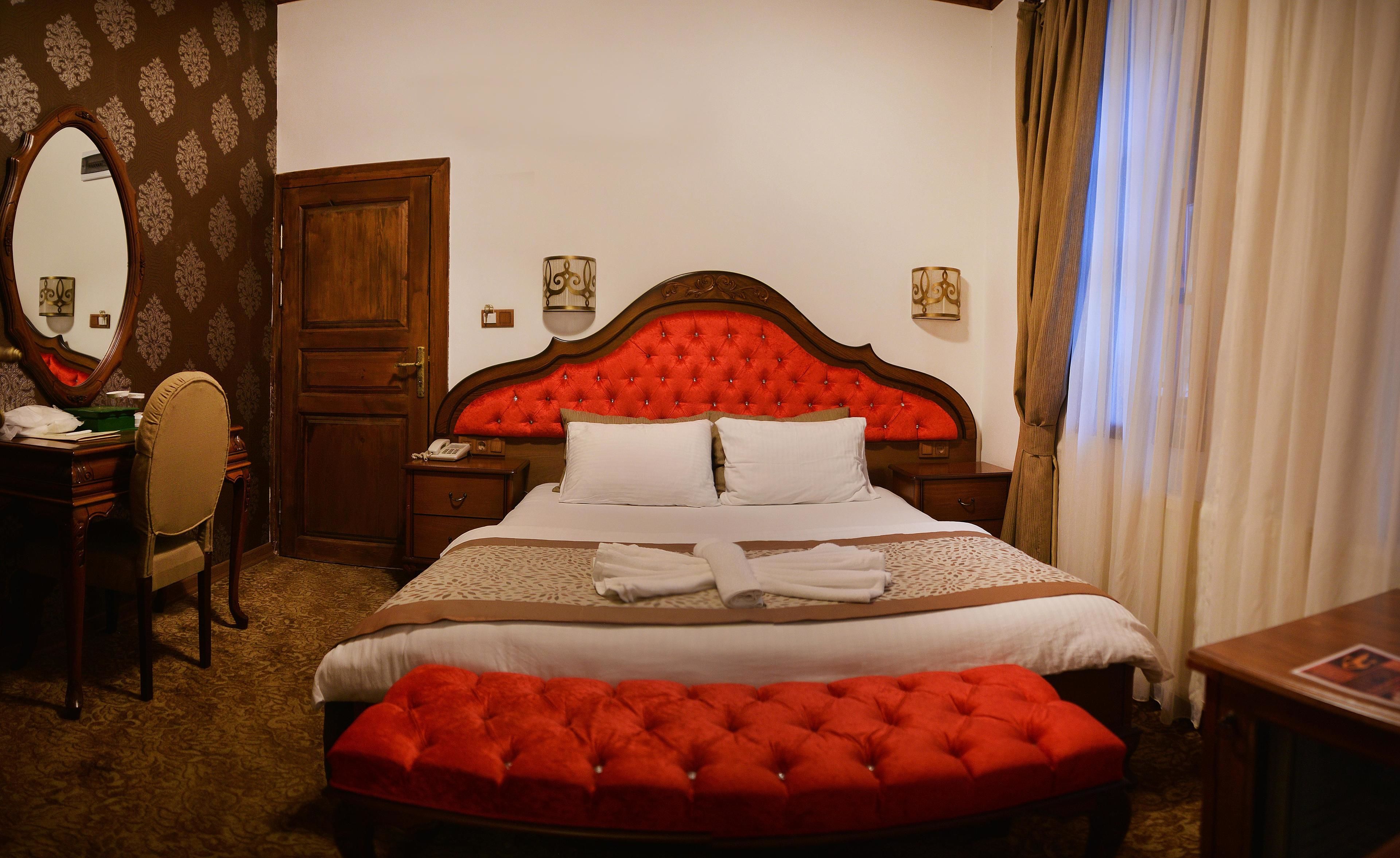 A hotel room with a large bed, a red tufted headboard, a wooden desk with a telephone, a chair, and a large mirror.