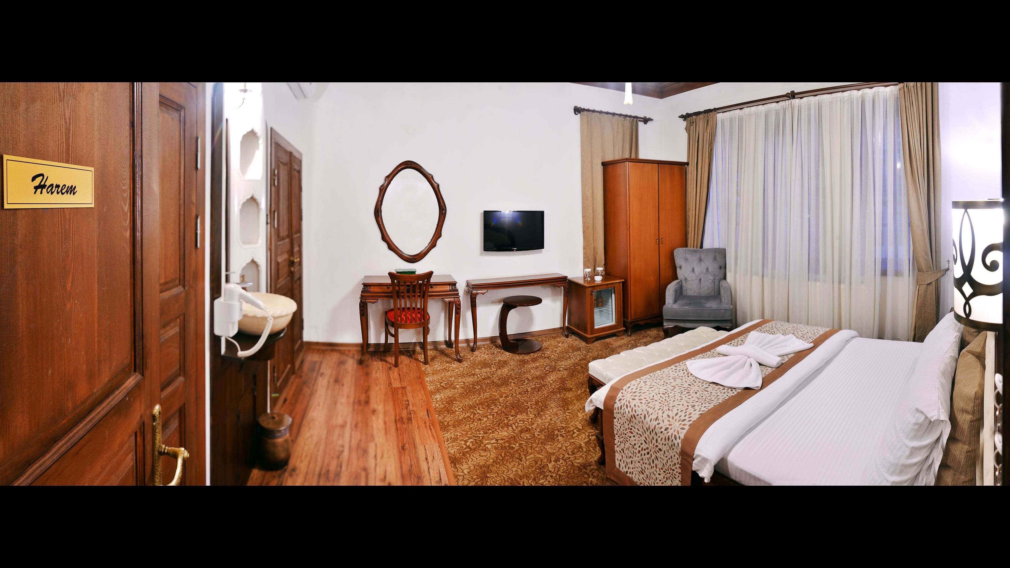 Une chambre d’hôtel avec une porte en bois, un bureau avec une chaise, une télévision, un lit avec des serviettes et une fenêtre avec des rideaux.
