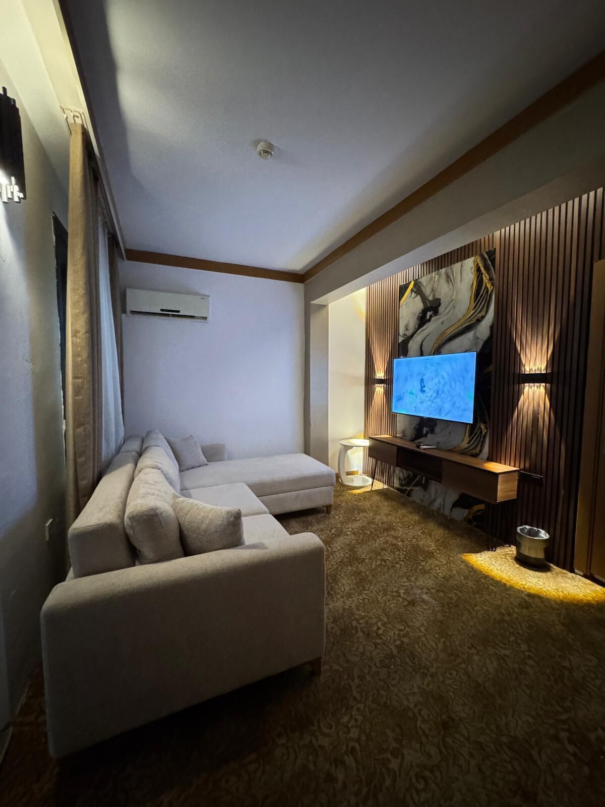 A modern living room with a sectional sofa, a flat-screen TV mounted on a wooden wall, and a small side table.