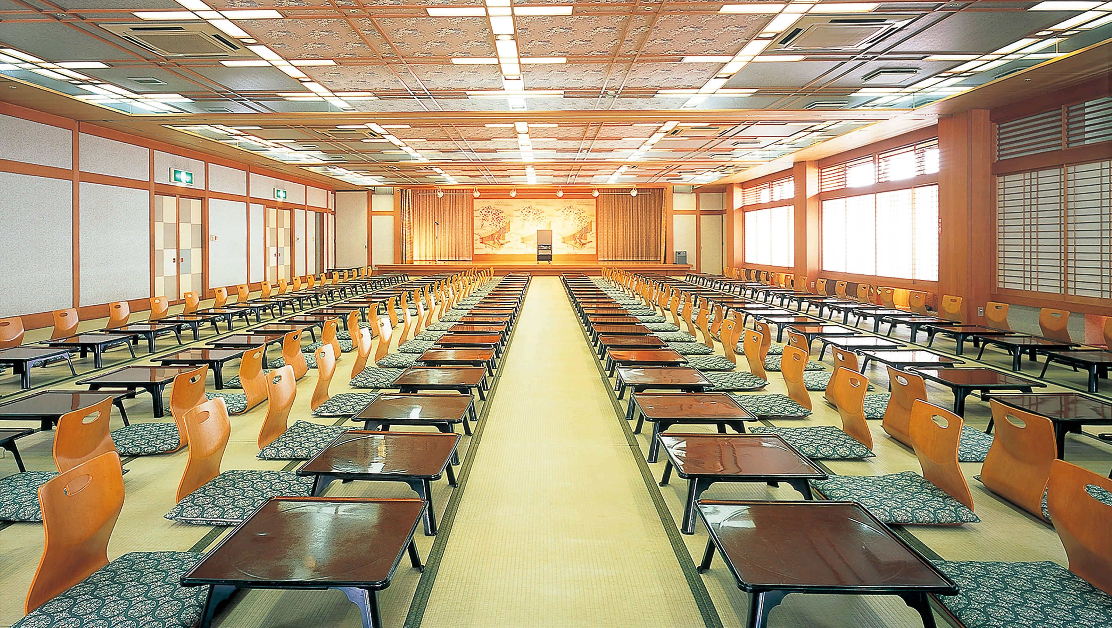Un salón de conferencias con filas de mesas y sillas, suelo alfombrado y un techo al estilo japonés con vigas de madera y farolillos de papel.