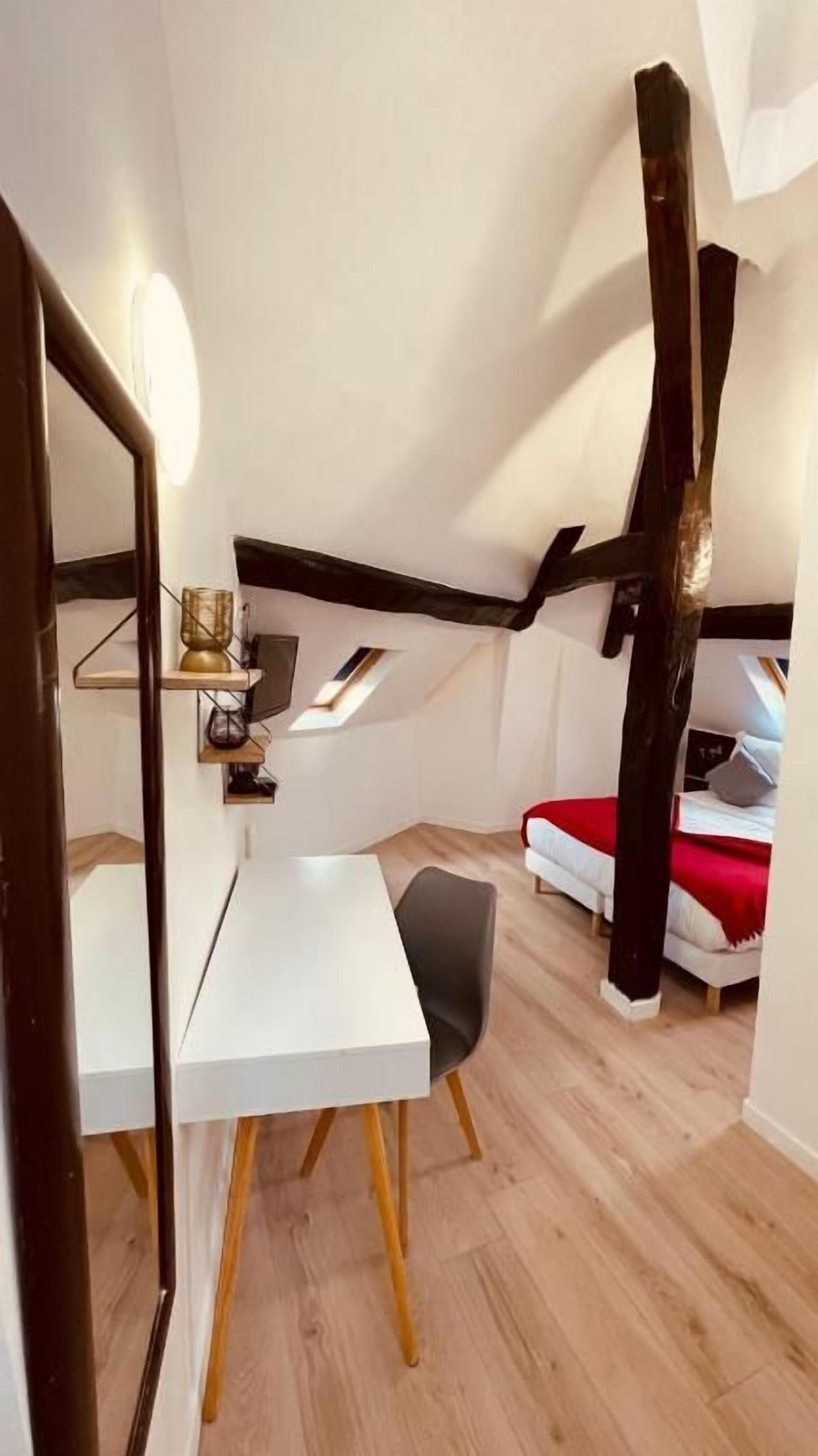 A small, compact room with a white desk, a chair, a bed with a red blanket, and a sloped ceiling with wooden beams.