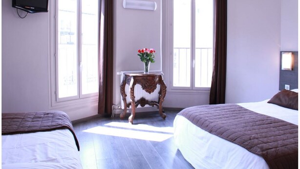 A hotel room with two beds, a small table with a vase of flowers, a flat-screen TV mounted on the wall, and large windows with curtains.