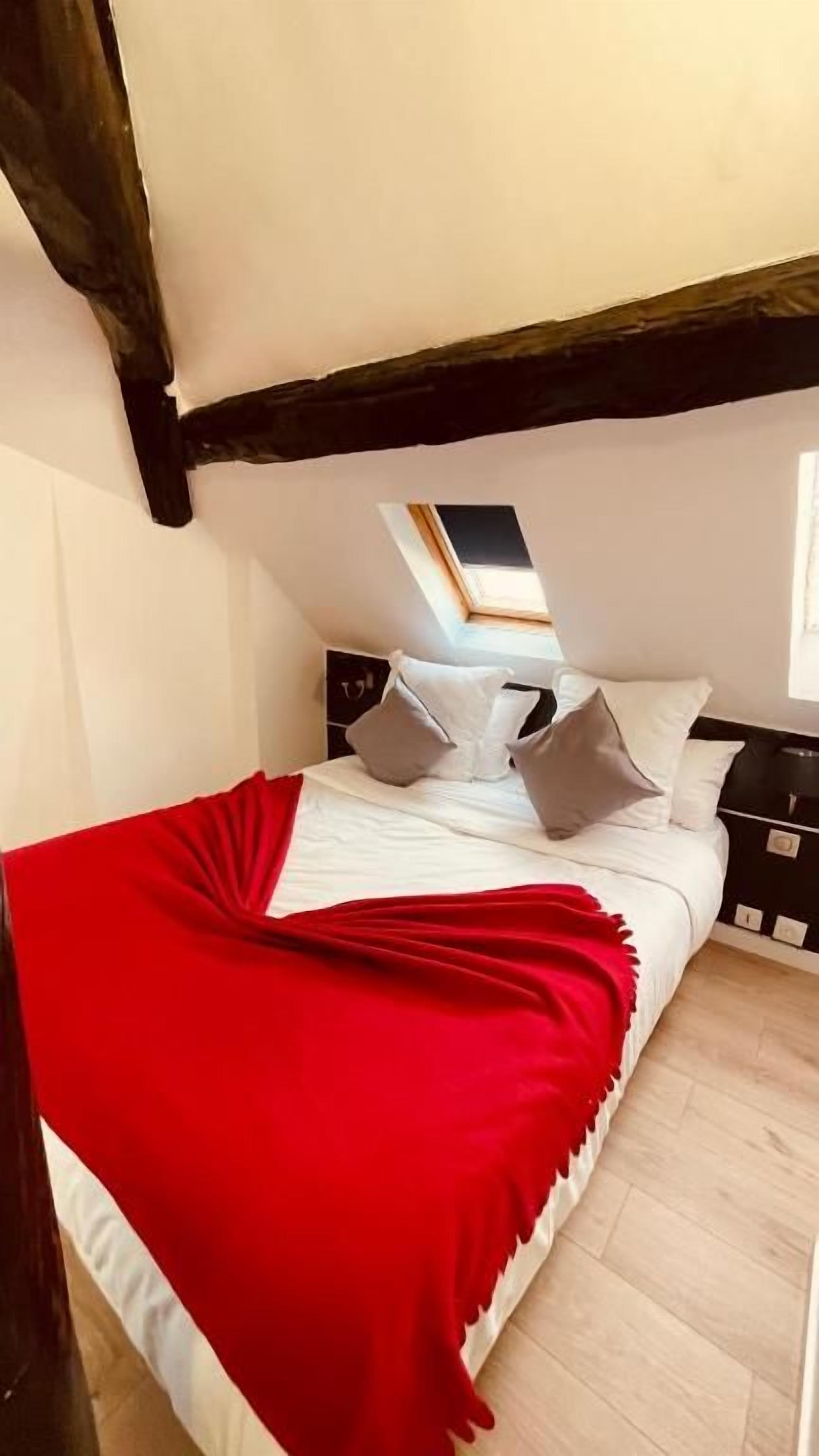 A bedroom with a sloped ceiling, a skylight, a bed with white bedding and a red throw, and a nightstand with a lamp.