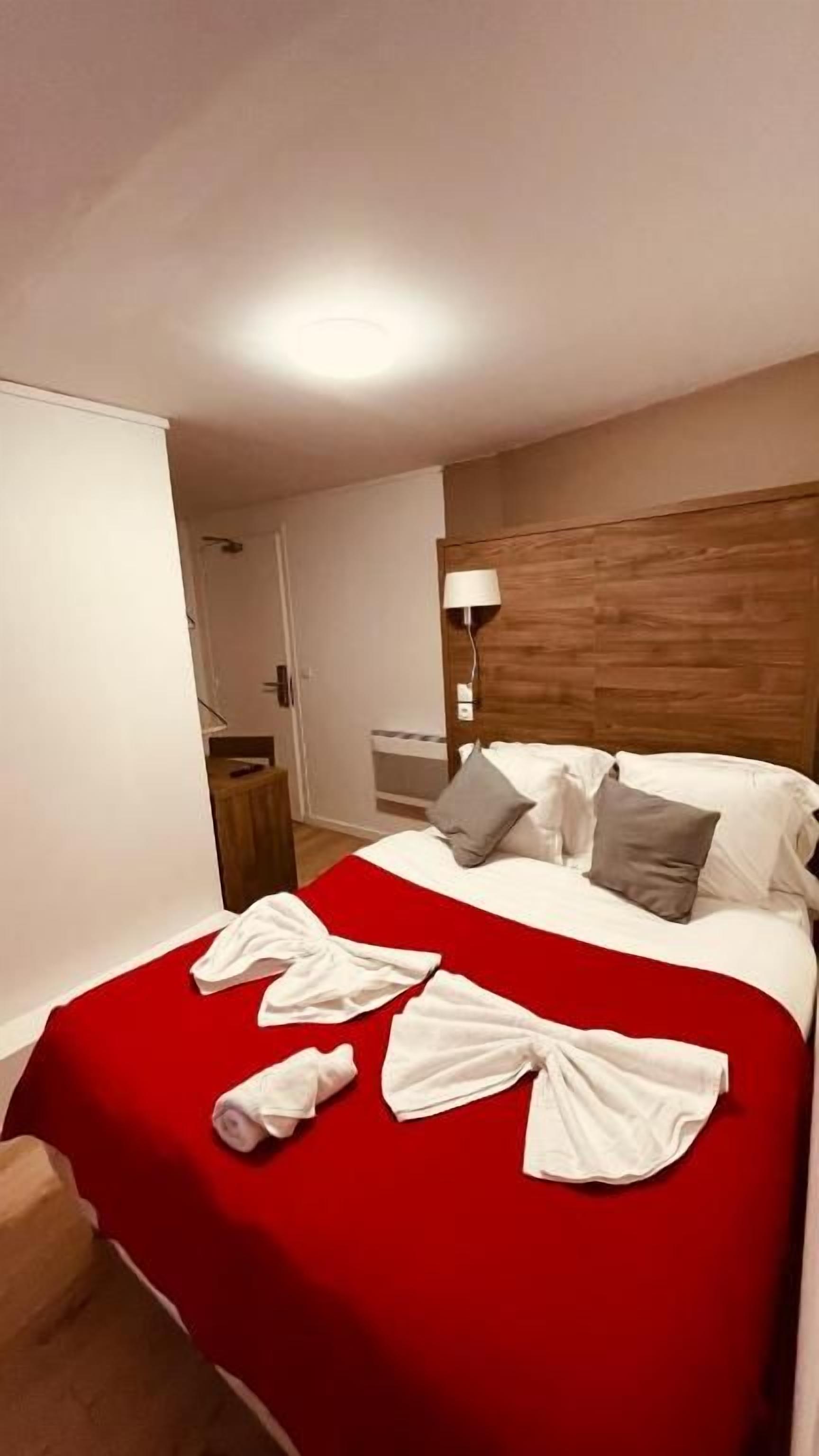 A hotel room with a bed, a red bedspread, white towels, a wooden headboard, and a nightstand with a lamp.