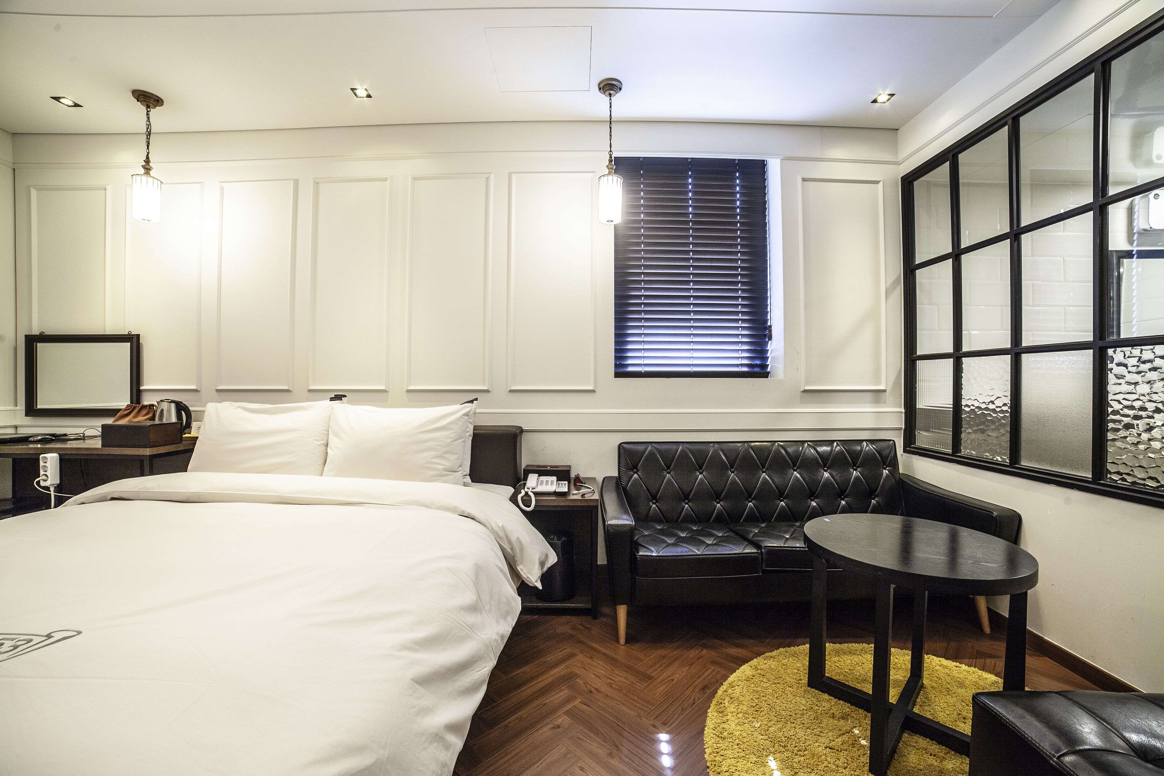 A hotel room with a bed, a black leather sofa, a small round table, a nightstand, and a window with blinds.