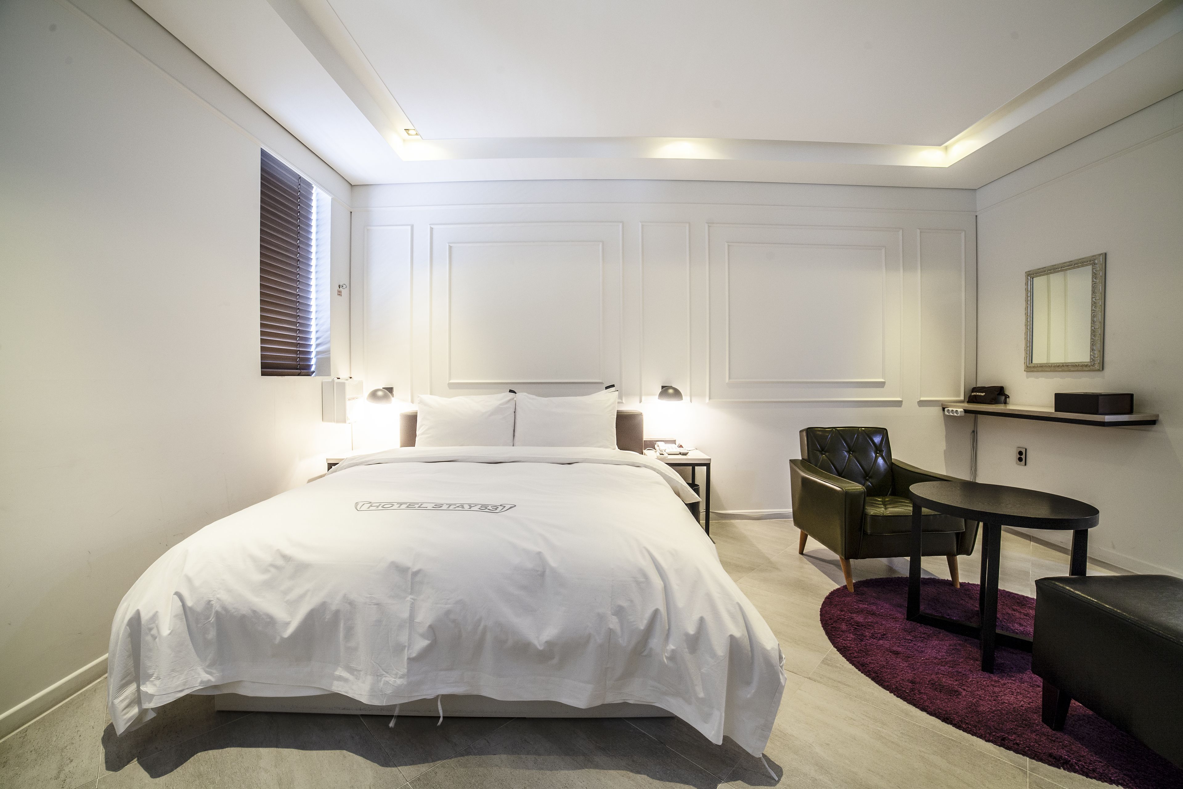 A modern hotel room with a large bed, bedside tables, a chair, a small table, and a mirror on the wall.
