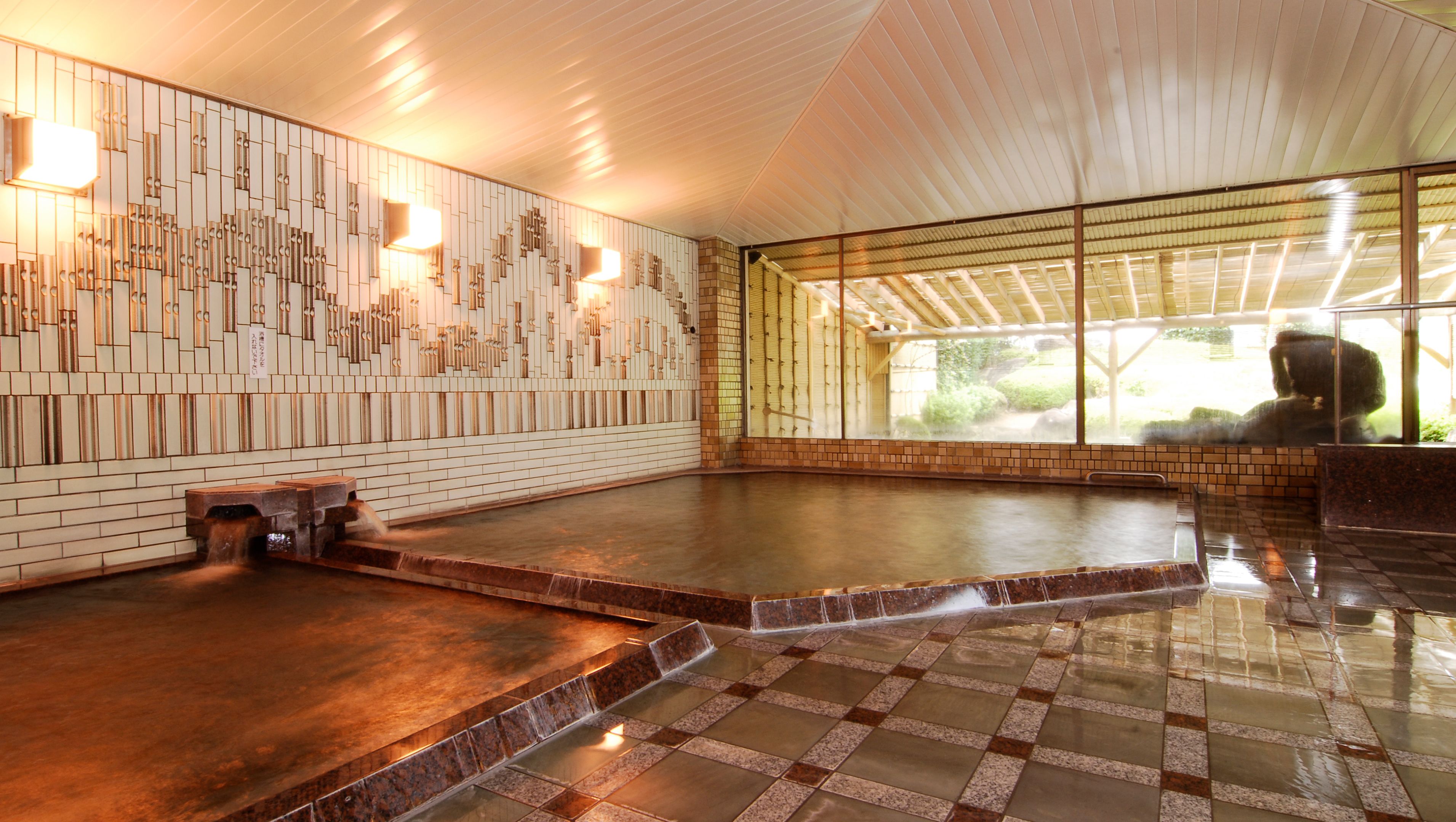 A spacious indoor area with a large, open floor plan, featuring a wall with a unique geometric pattern and a large window providing a view of the outdoors.