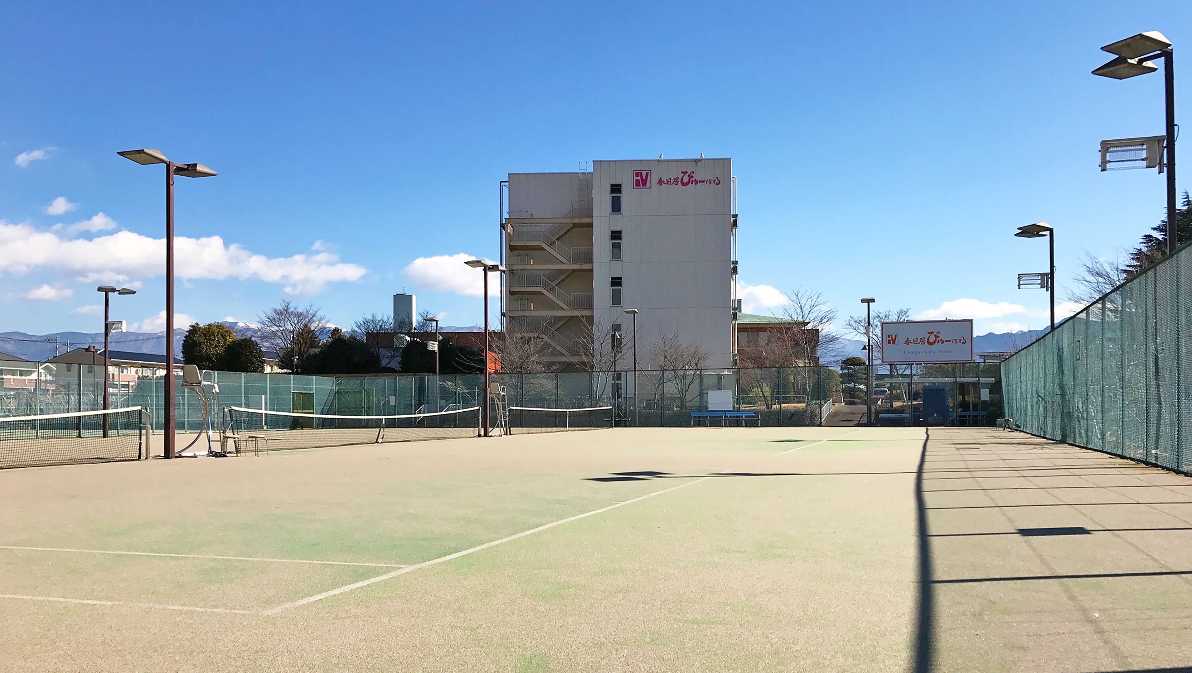 An outdoor tennis court with a net, surrounded by a fence, and a building with a sign in the background.
