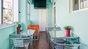 A narrow corridor with teal walls, a white door, and a TV mounted on the wall. There are tables and chairs along the corridor, and potted plants on the tables. - Hostel Seven Hills (Lisbon)