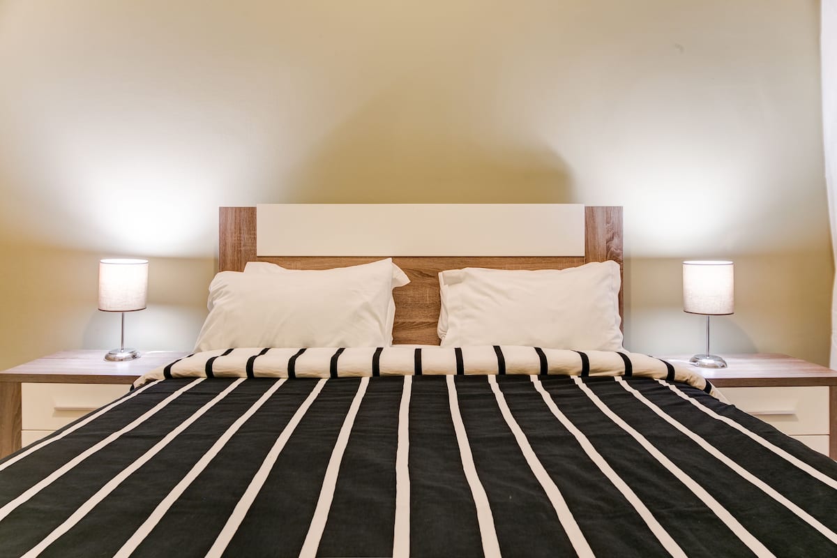 Ein ordentlich bezogenes Bett mit einem gestreiften Bettbezug, zwei Nachttischlampen und einem hölzernen Kopfteil.