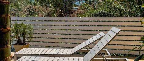 Una tumbona con estructura metálica y tiras de madera, ubicada en un patio de concreto con valla de madera y palmeras al fondo.