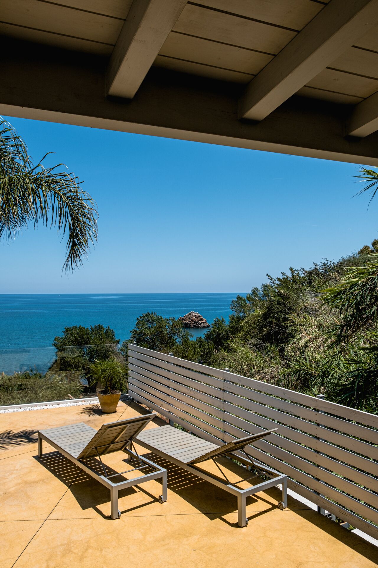 Ein Balkon mit zwei Liegestühlen und einem Blumentopf, mit Blick auf das klare blaue Meer und einen Felsen.