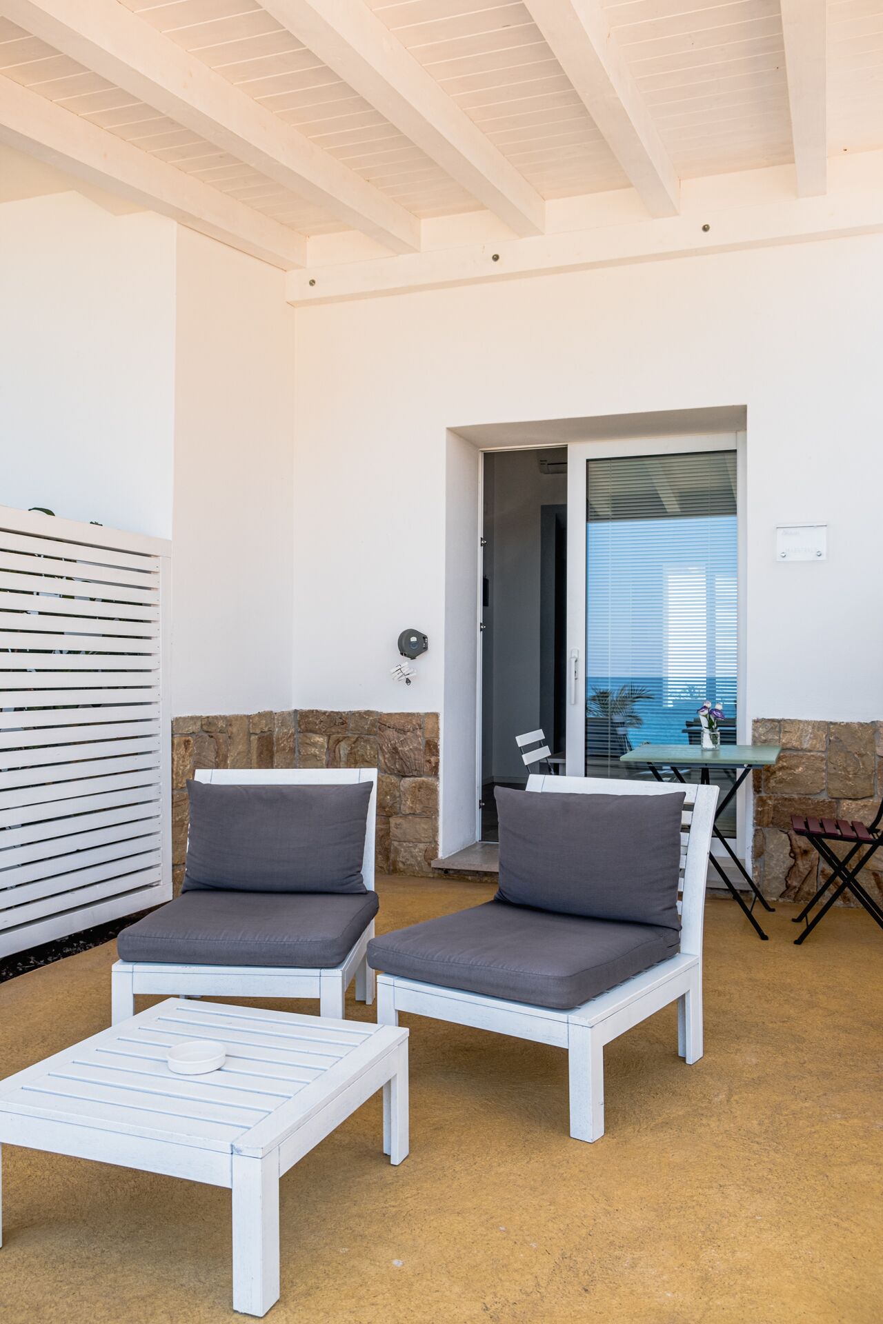 Un patio soleado con dos sillas blancas acolchadas y una mesita, un muro de piedra y vistas a través de una puerta de cristal.