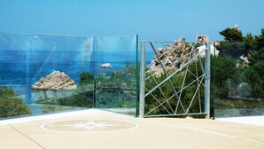 A glass enclosure with a metal frame, a paved area with a compass symbol, and a view of the sea and rocky landscape. - A Tonnara (Cefalù)