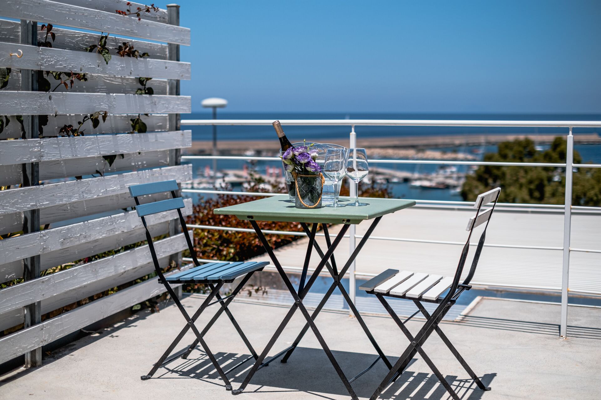 A table with a bottle of champagne, two wine glasses, and a vase with flowers on a balcony overlooking a marina.