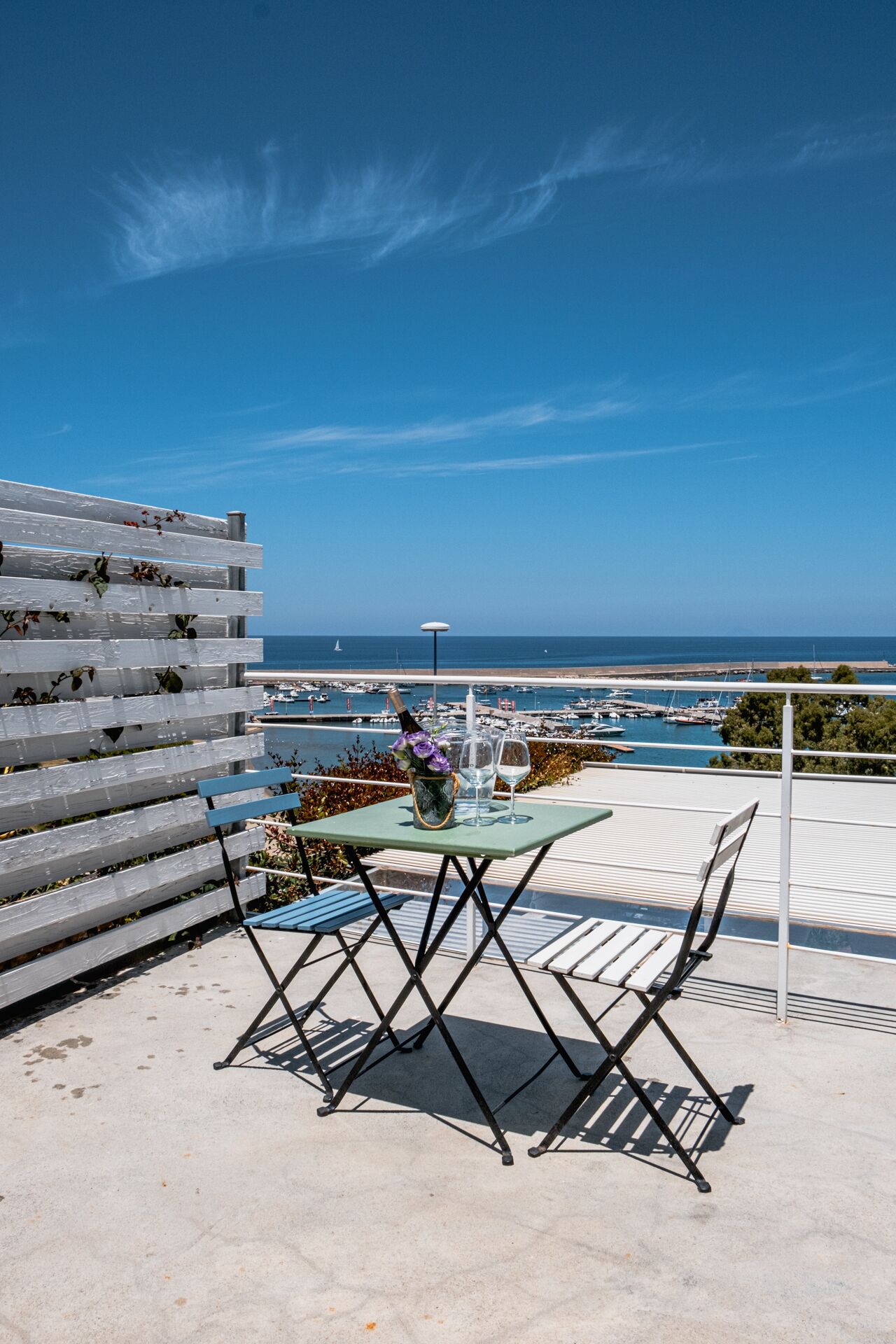 Un espacio al aire libre con mesa y sillas, con vista a un puerto deportivo con barcos y un cielo azul despejado.