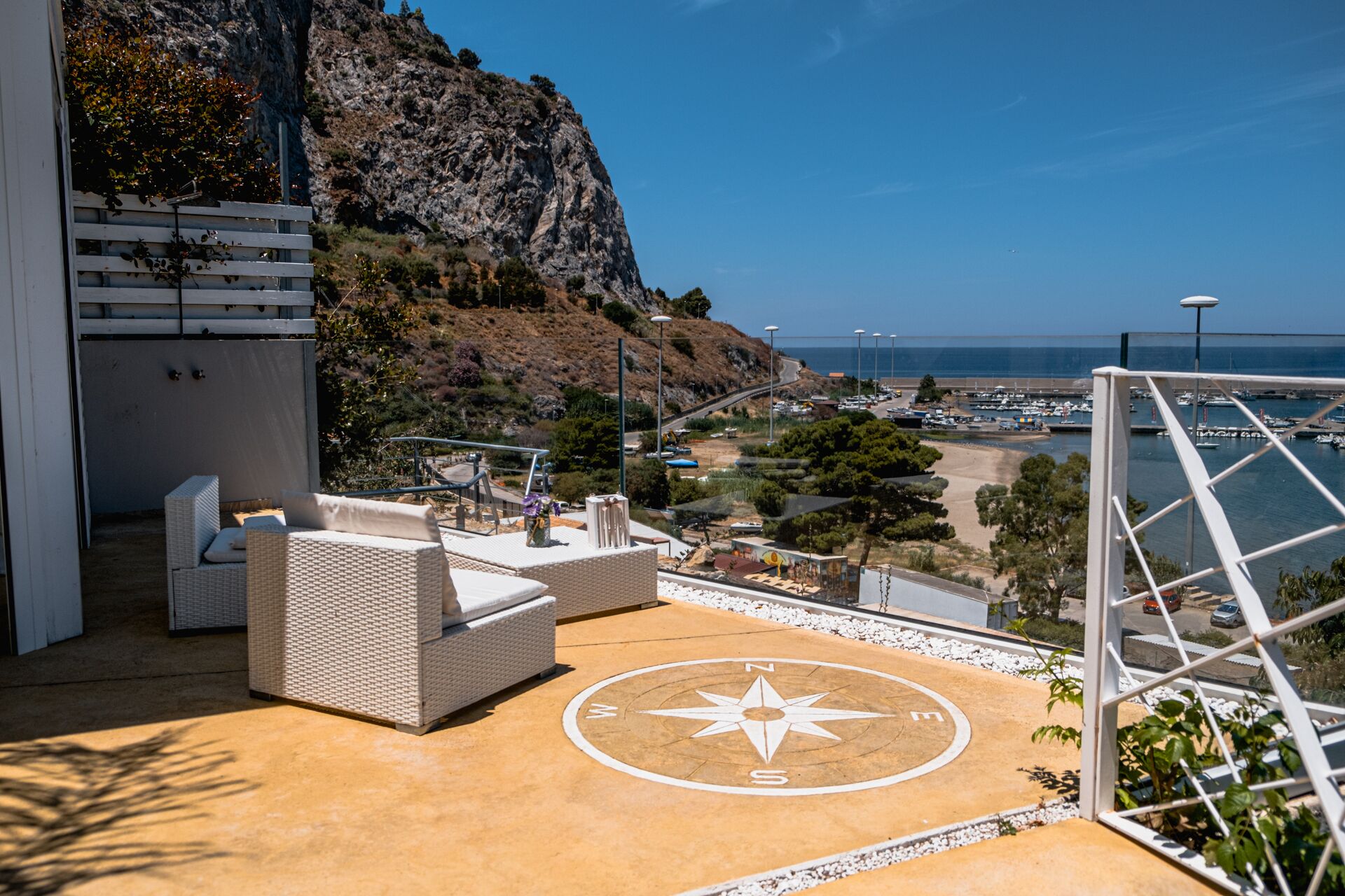 A rooftop terrace with outdoor seating, a compass design on the floor, and a view of the coastline and marina.