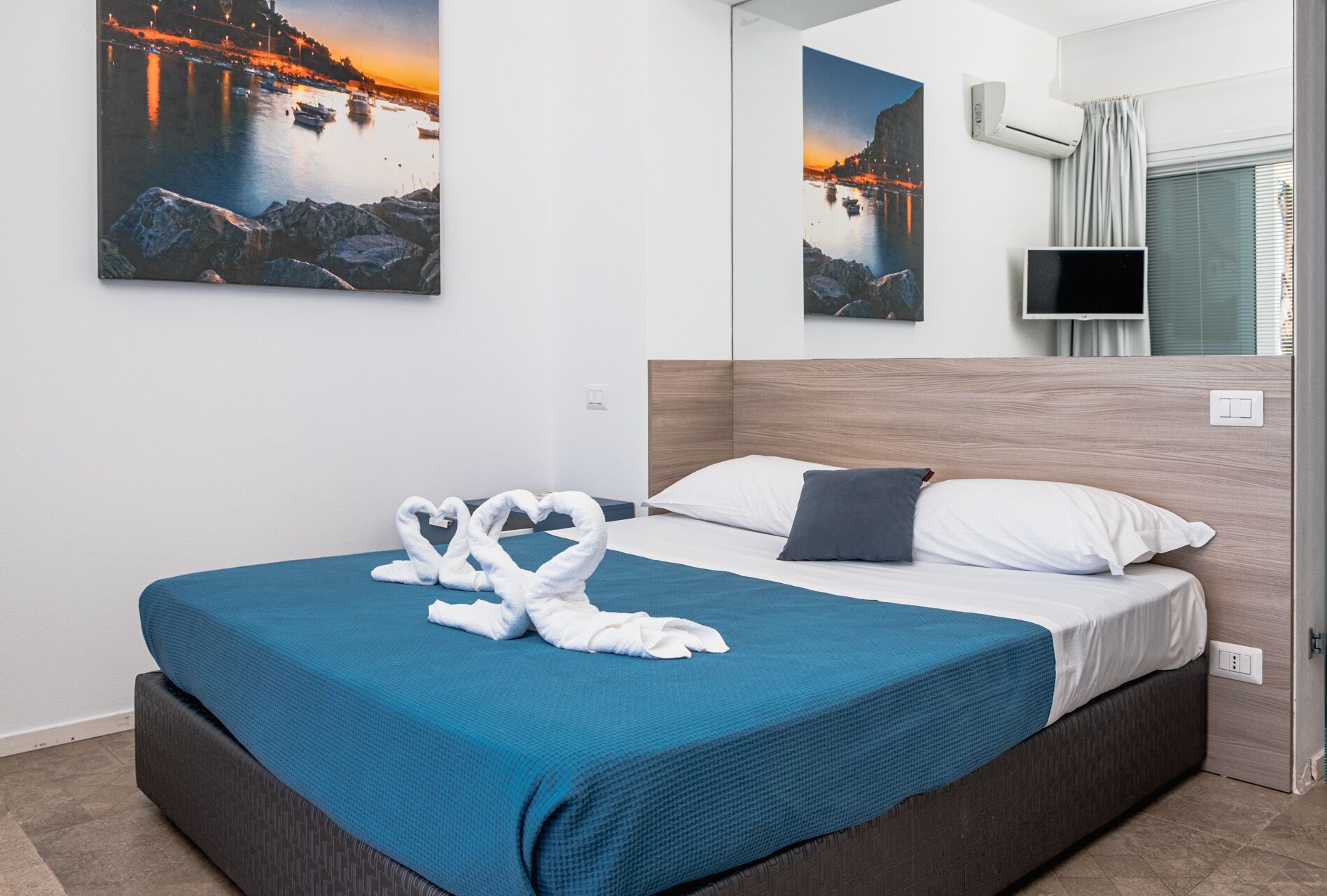 Quarto de hotel com uma cama, uma televisão e duas pinturas de uma paisagem costeira.