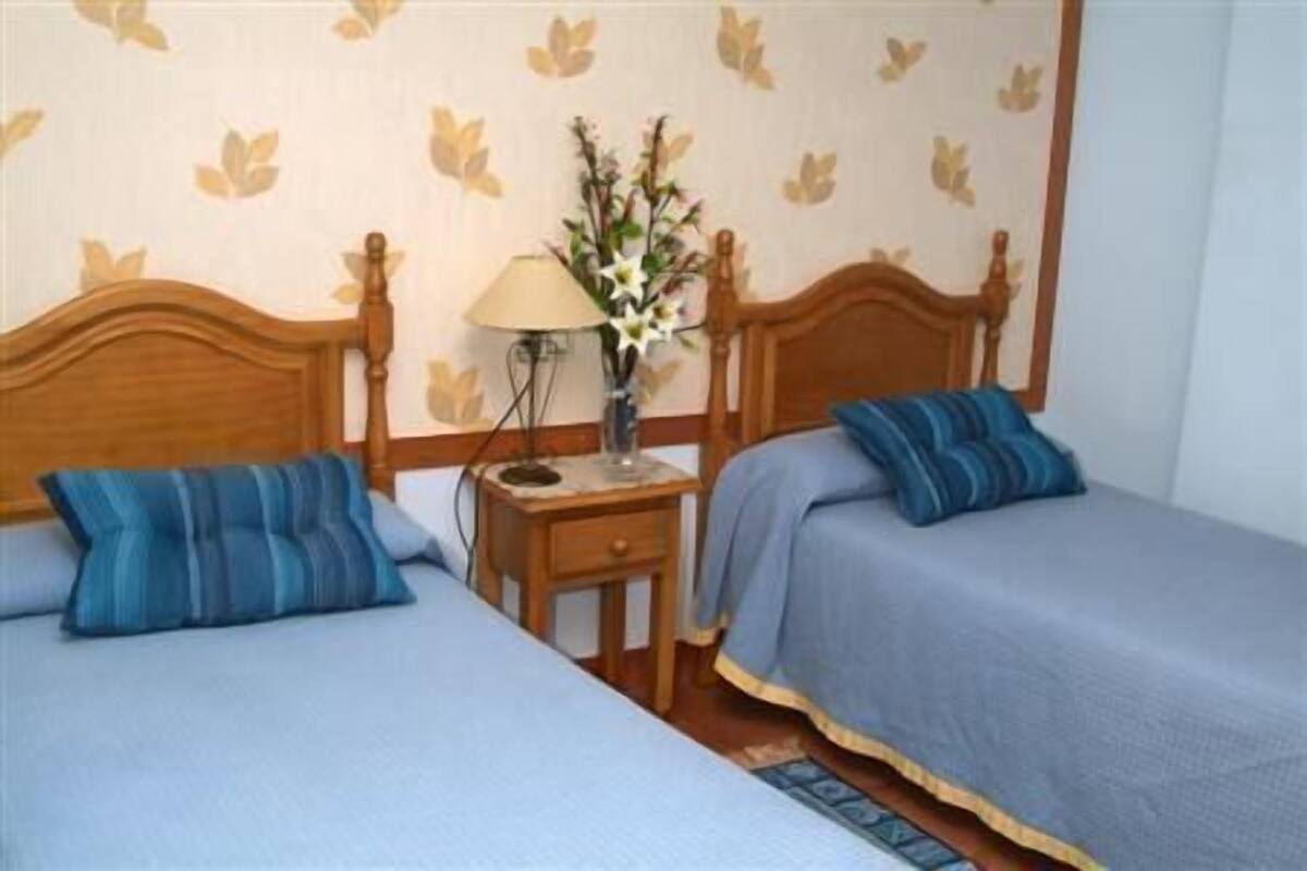 A room with two single beds, a nightstand with a lamp and a vase of flowers, and a patterned wallpaper.
