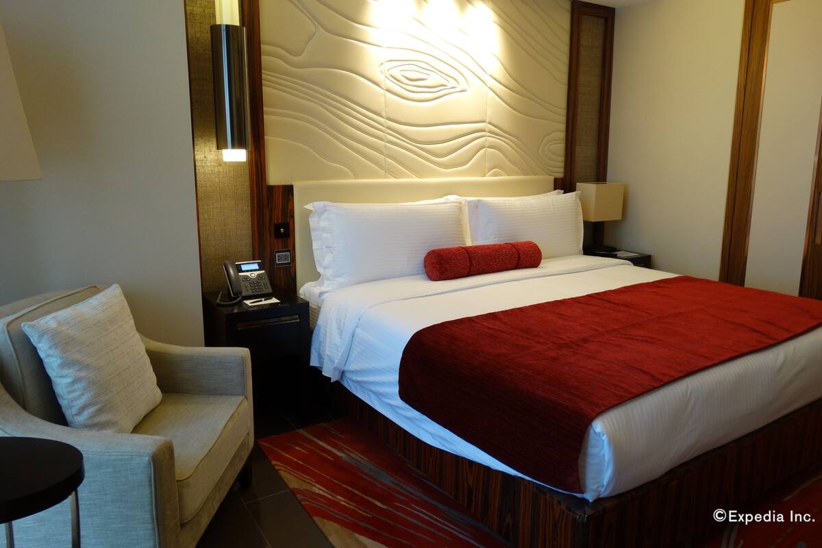 Habitación de hotel con cama, mesita de noche, teléfono, lámpara, silla y pared con diseño texturizado.