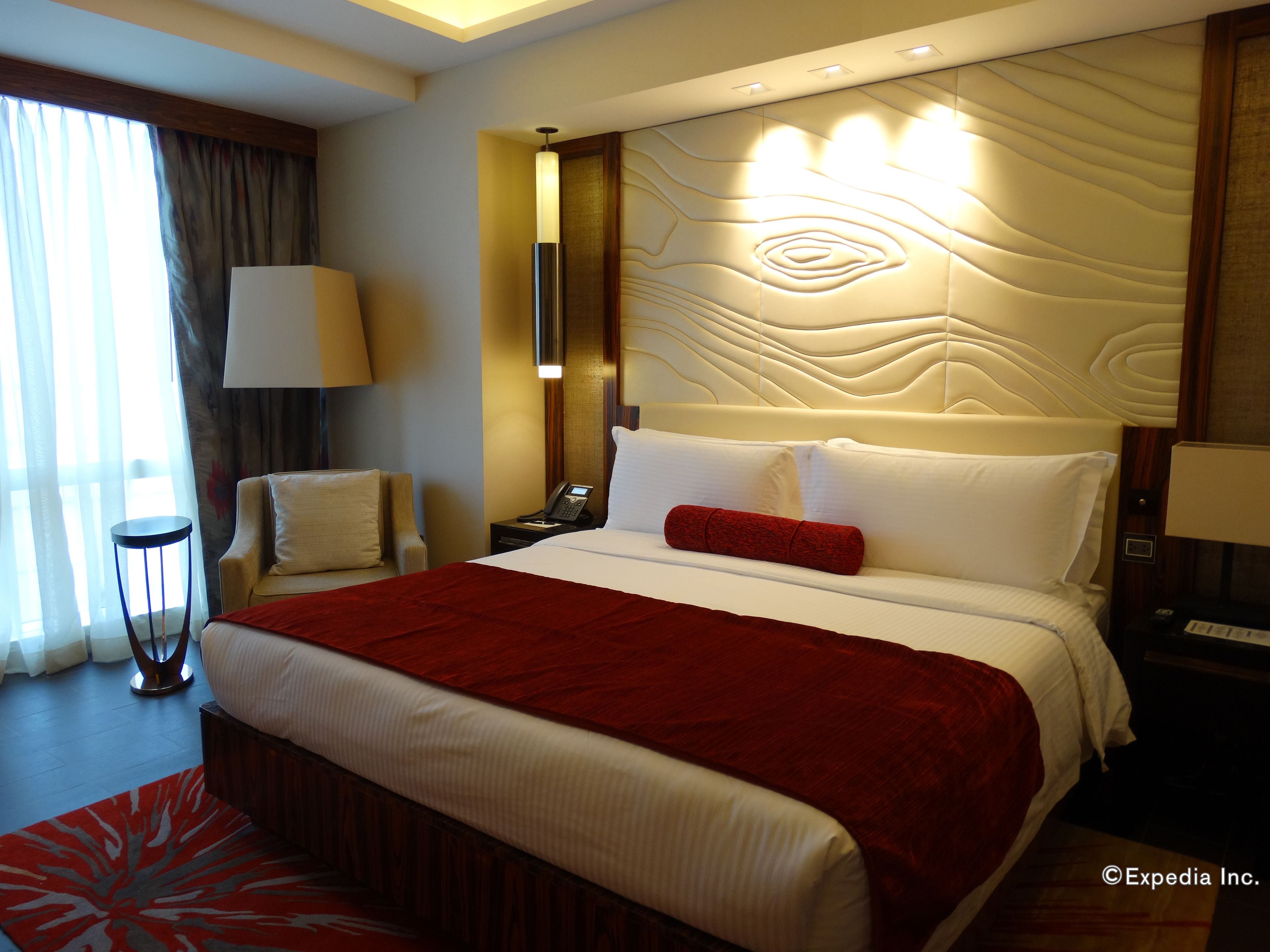 A hotel room with a large bed, a bedside table with a lamp, a chair, and a view of the outdoors through a window.