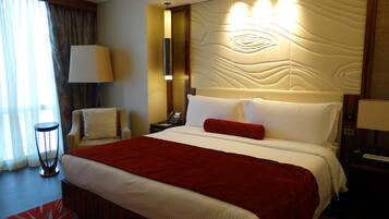 A hotel room with a large bed, a bedside table with a lamp, a chair, and a view of the outdoors through a window.