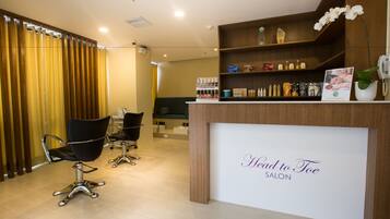 Un interior de salón de belleza con una recepción, dos sillas negras para peluquería y varios productos para el cuidado del cabello en estantes.