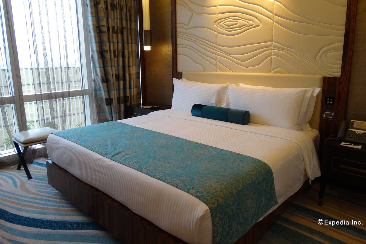 Una habitación de hotel con una cama grande, cabecera de madera, mesita de noche, teléfono y ventana con cortinas.