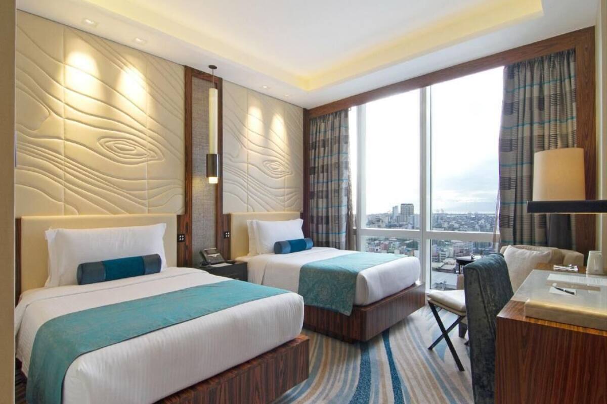 Een hotelkamer met twee bedden, een bureau, een stoel en een groot raam met uitzicht op de stad.