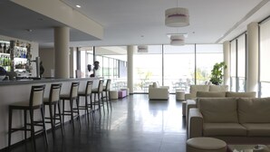 A modern bar area with a long counter, bar stools, and a variety of bottles. There is a seating area with a sofa and chairs, and large windows providing a view of the outdoors. - Monte Lírio Hotel (Espinho)
