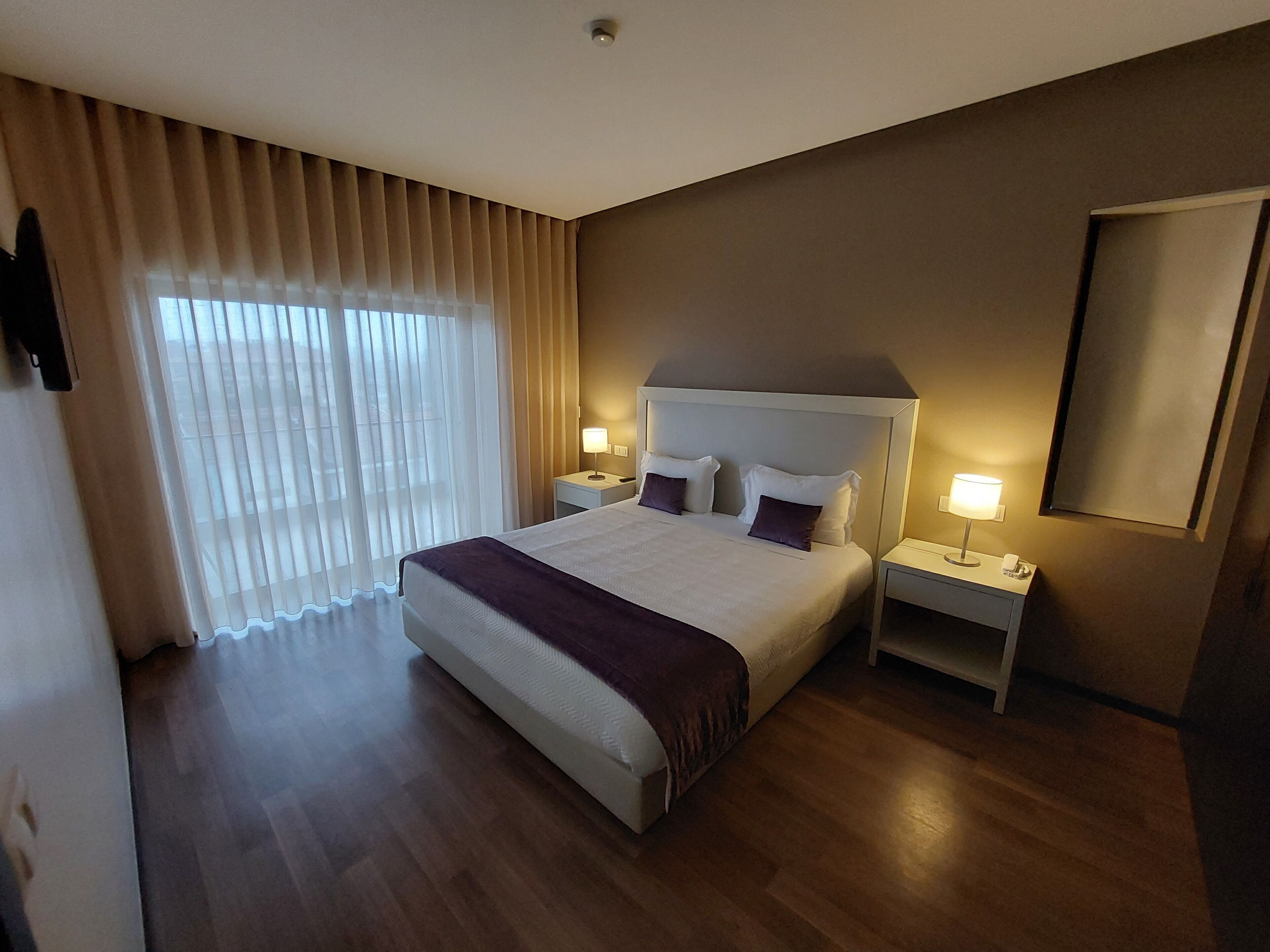 A hotel room with a large bed, two bedside tables, a nightstand, a mirror, and a window with curtains.