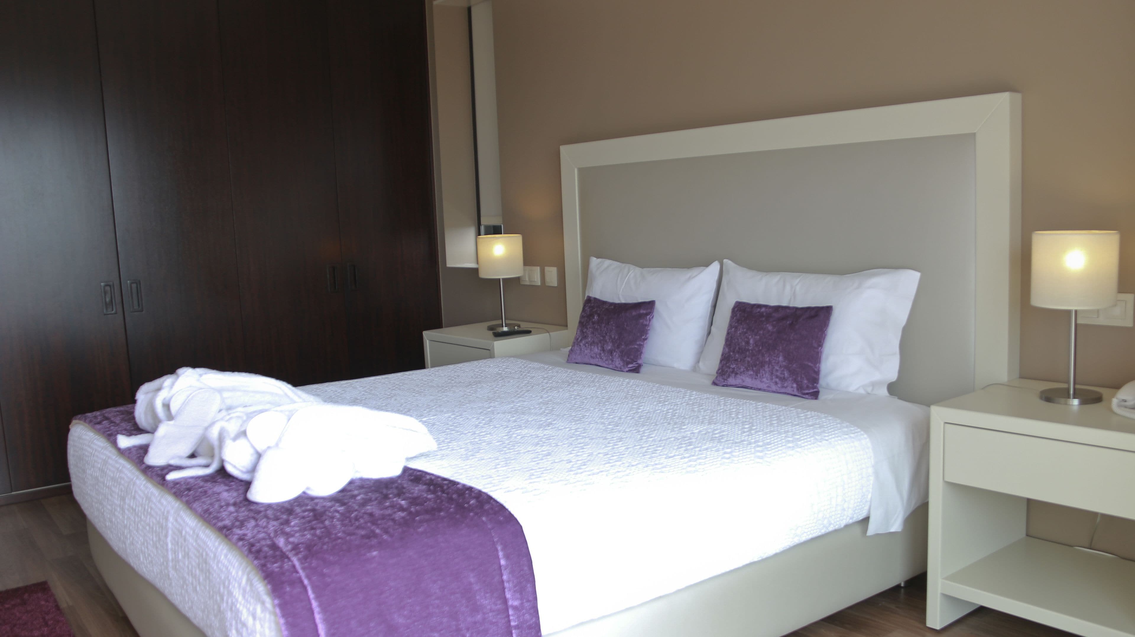 A neatly made bed with a white and purple blanket, two white pillows, and a purple throw. There are two bedside tables with lamps, a wooden wardrobe, and a light-colored wall.