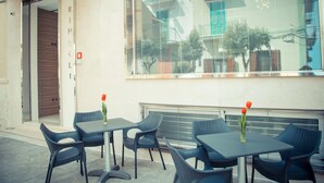 An outdoor seating area with tables and chairs, a glass door entrance, and a large window reflecting the building's exterior. - Hotel Ristorante Rinelli (Margherita di Savoia)