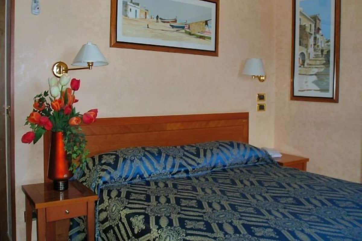 Une chambre d’hôtel avec une tête de lit en bois, une couvre-lit à motifs, un vase de fleurs, une lampe de chevet et deux tableaux encadrés au mur.