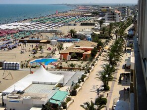Un hotel sul lungomare con vista spiaggia, piscina e una strada alberata di palme.