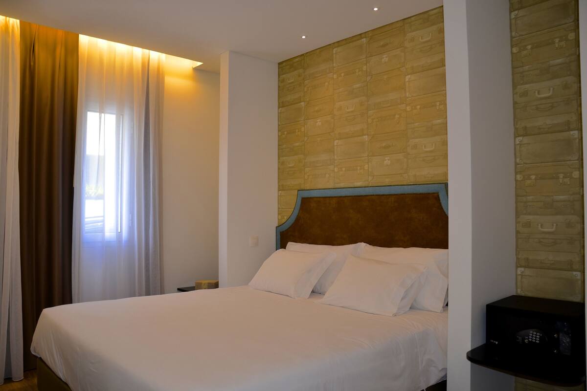 A hotel room with a large bed, a nightstand, a wall with a suitcase pattern, and a window with curtains.