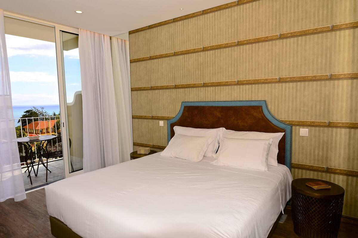 A hotel room with a large bed, a view of the ocean, a balcony with a table and chairs, and a wooden headboard.