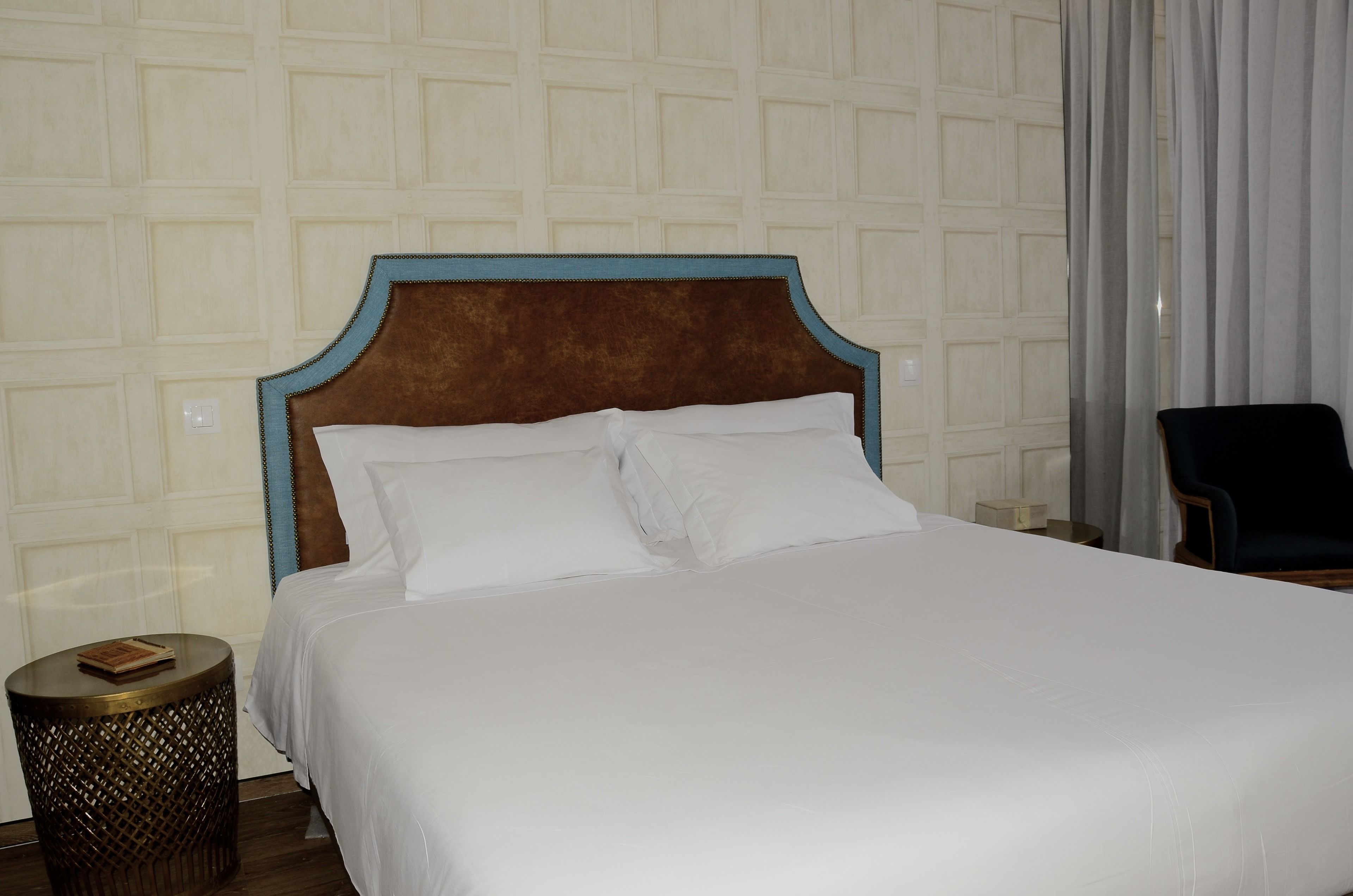 Una cama bien hecha con cabecera marrón, una mesita de madera redonda con un libro, y una silla en una habitación de hotel con pared estampada.