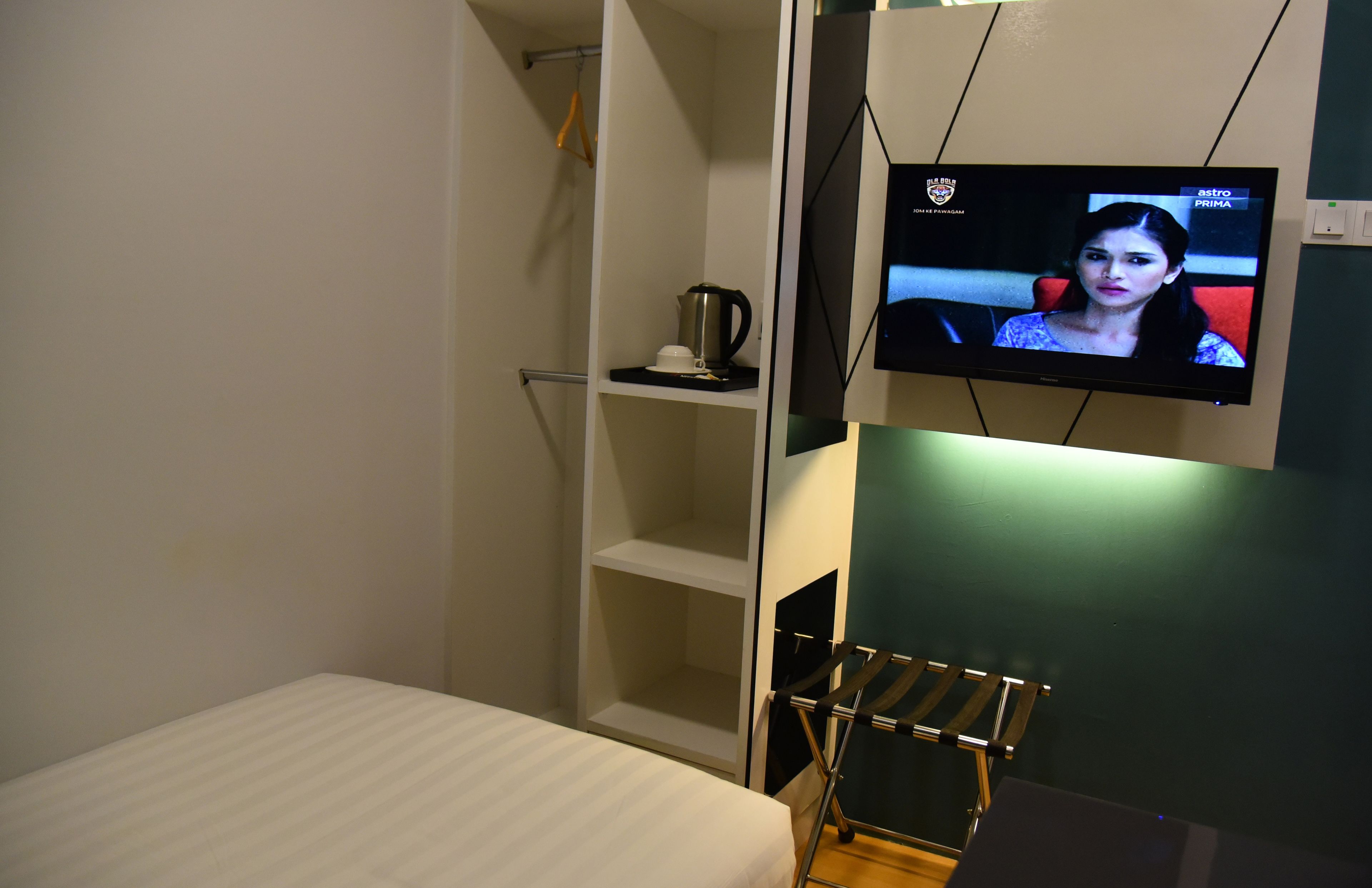 A hotel room with a bed, a shelf with a kettle, and a TV mounted on the wall.