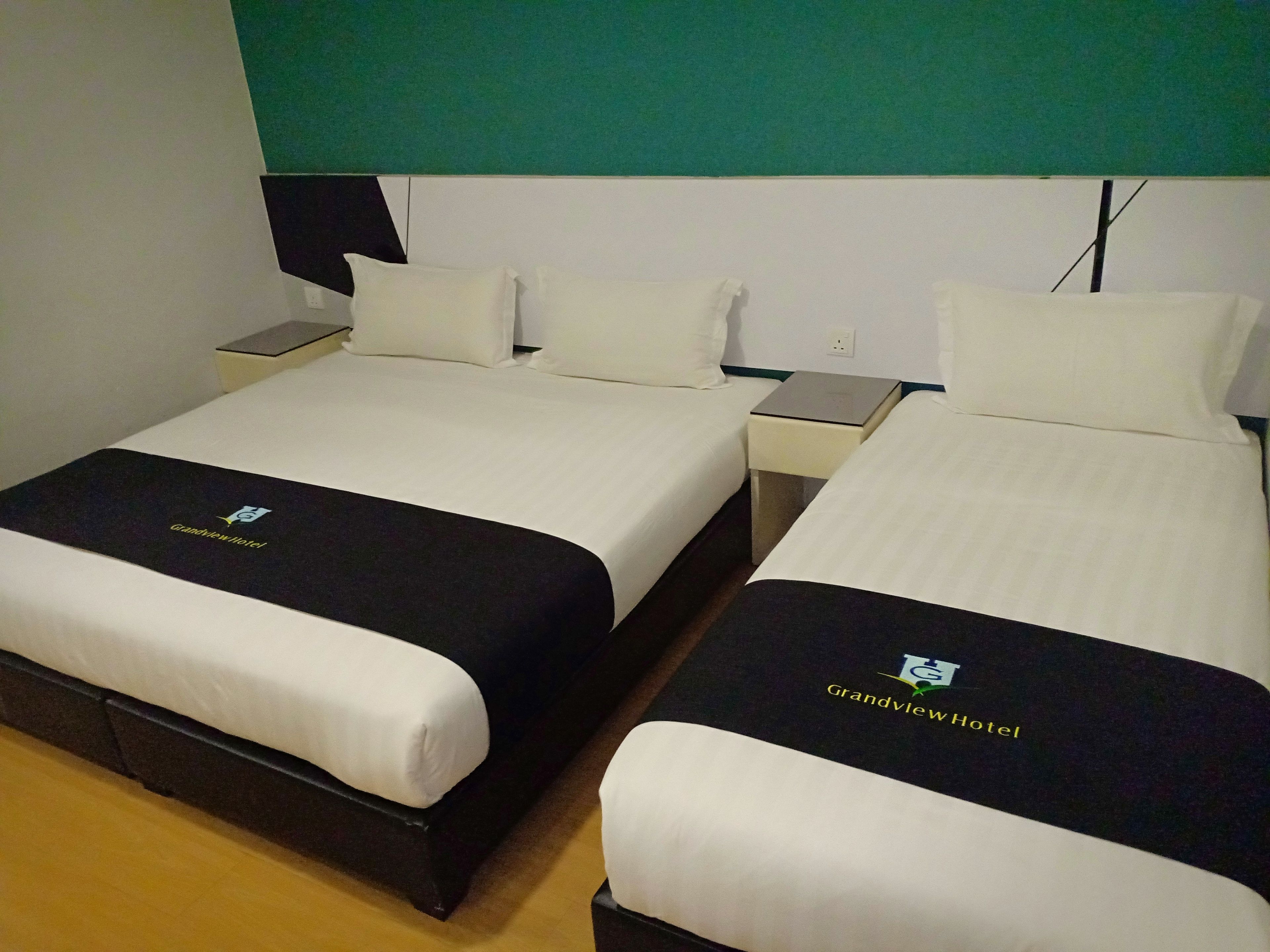A hotel room with two beds, each with white bedding and a black headboard. There is a nightstand with a lamp and a power outlet on the wall.