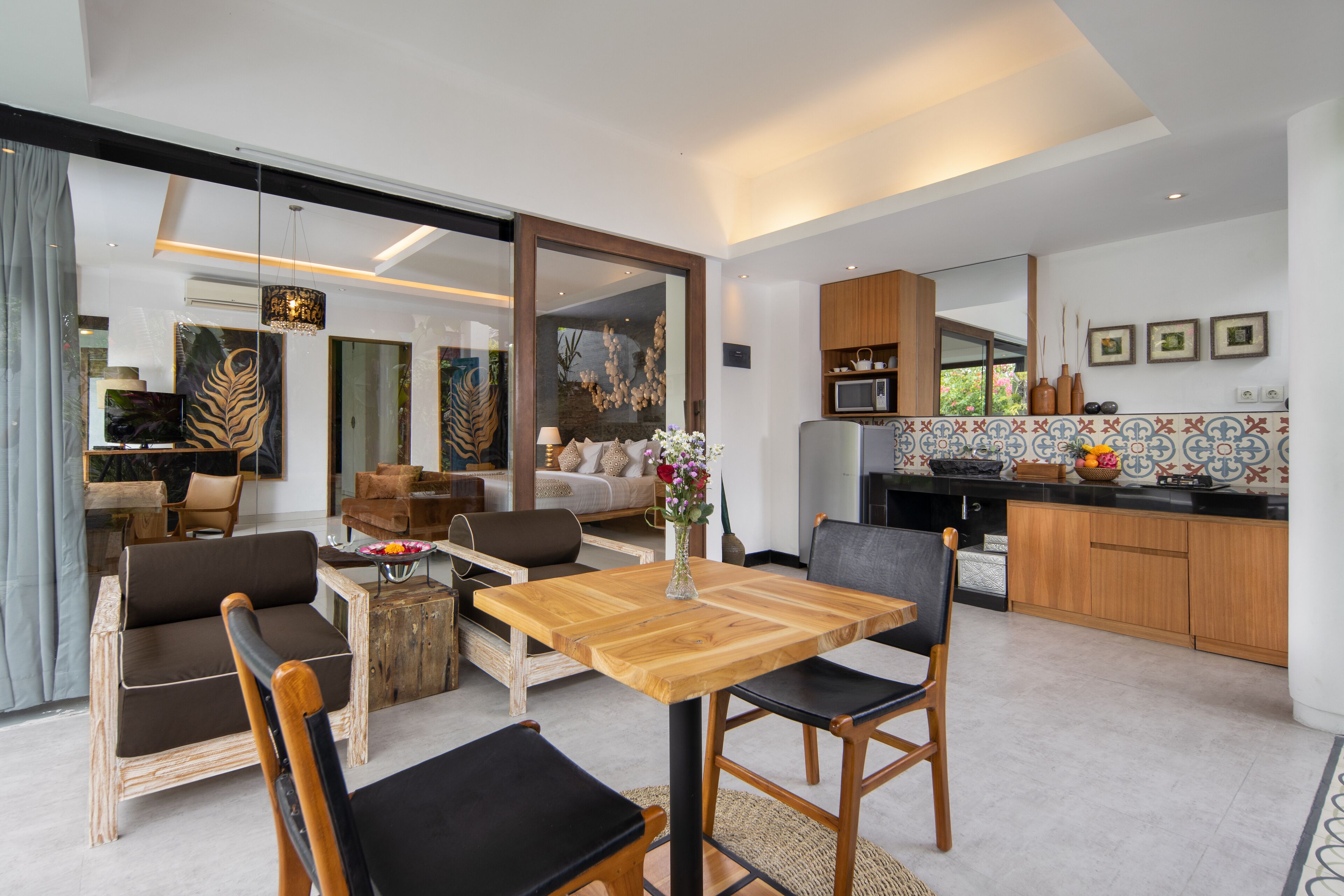 A modern living room with a wooden dining table, leather chairs, and a sofa. There is a kitchen area with wooden cabinets and a tiled backsplash.