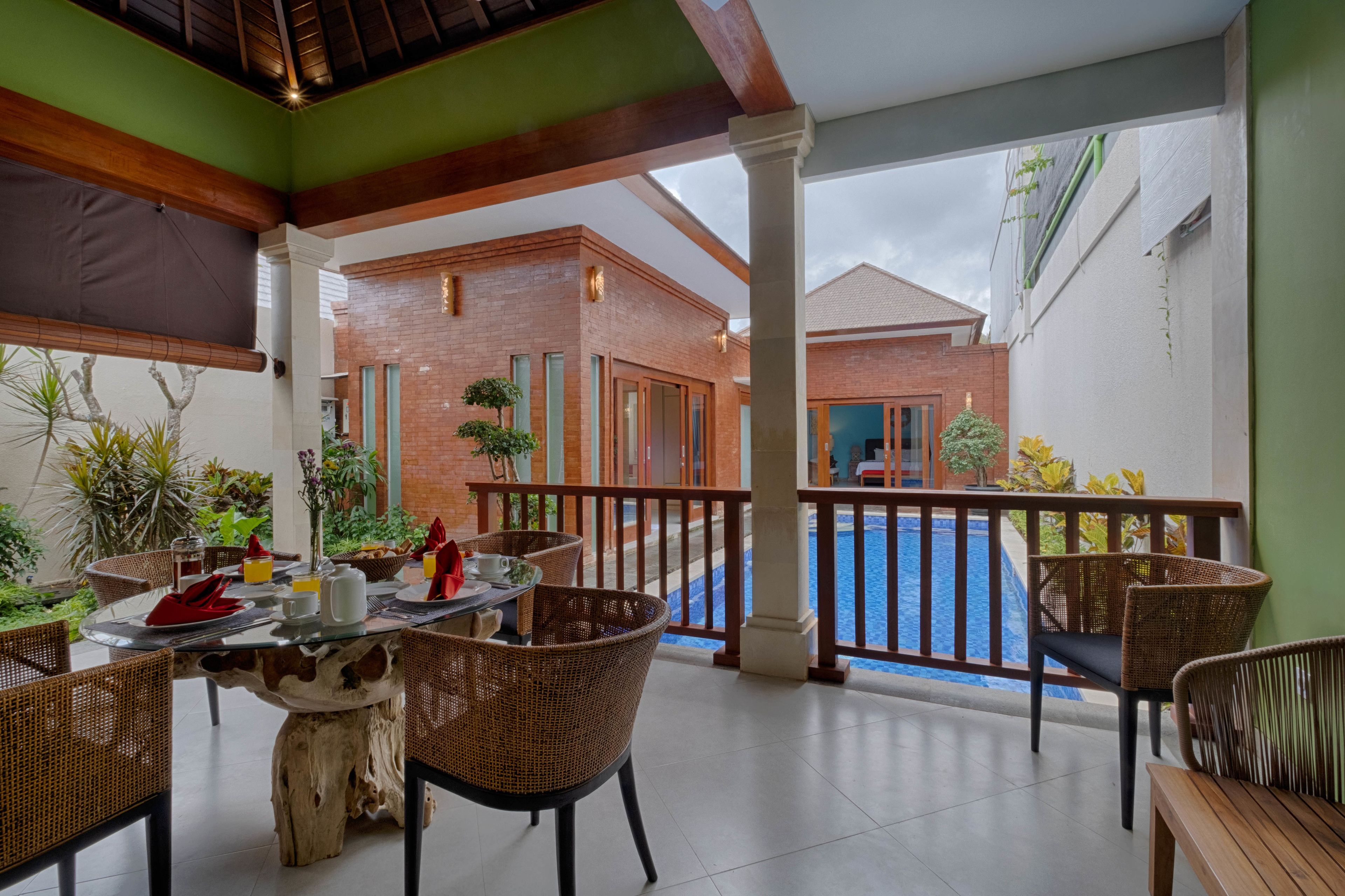 Un área de comedor al aire libre techada con sillas de mimbre, una mesa de vidrio y vistas a una piscina.