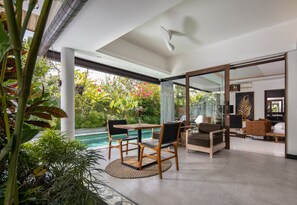 A modern living room with a pool, wooden furniture, and a glass door leading to a garden.