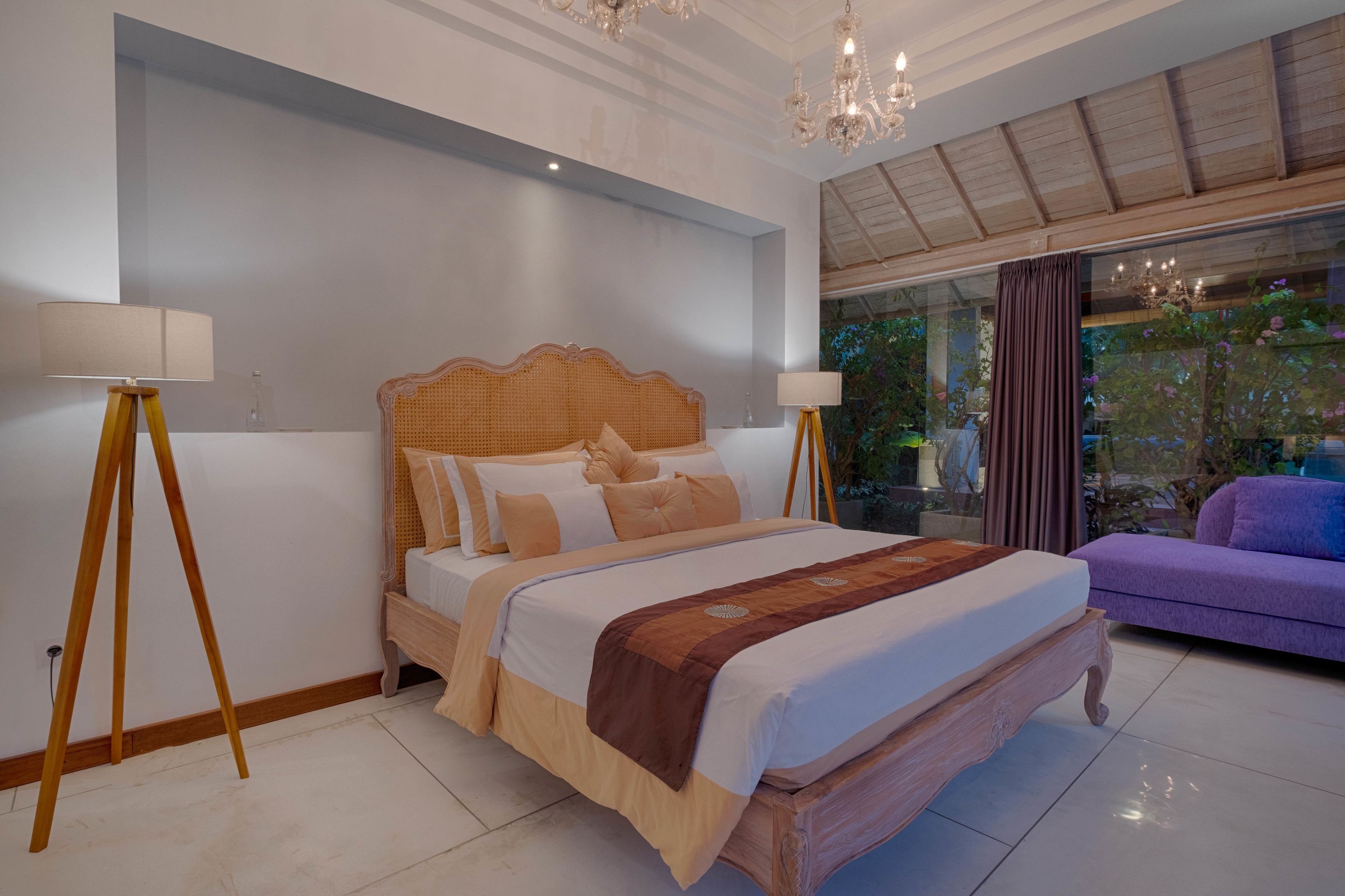 A bedroom with a large bed, two bedside lamps, a chandelier, and a view of greenery through a window.