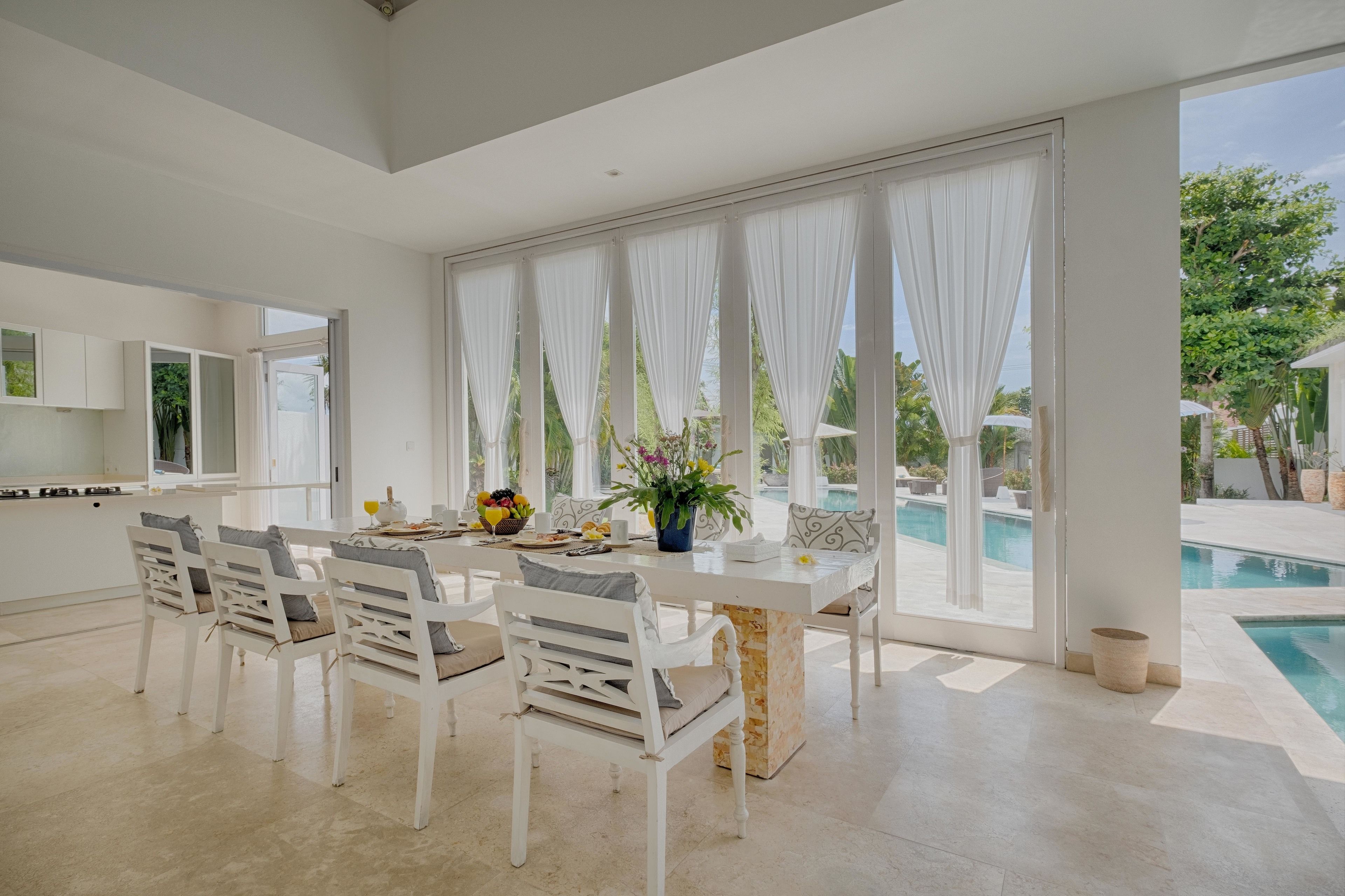 A modern kitchen with white cabinets, a dining area with white chairs, and a large window overlooking a pool.