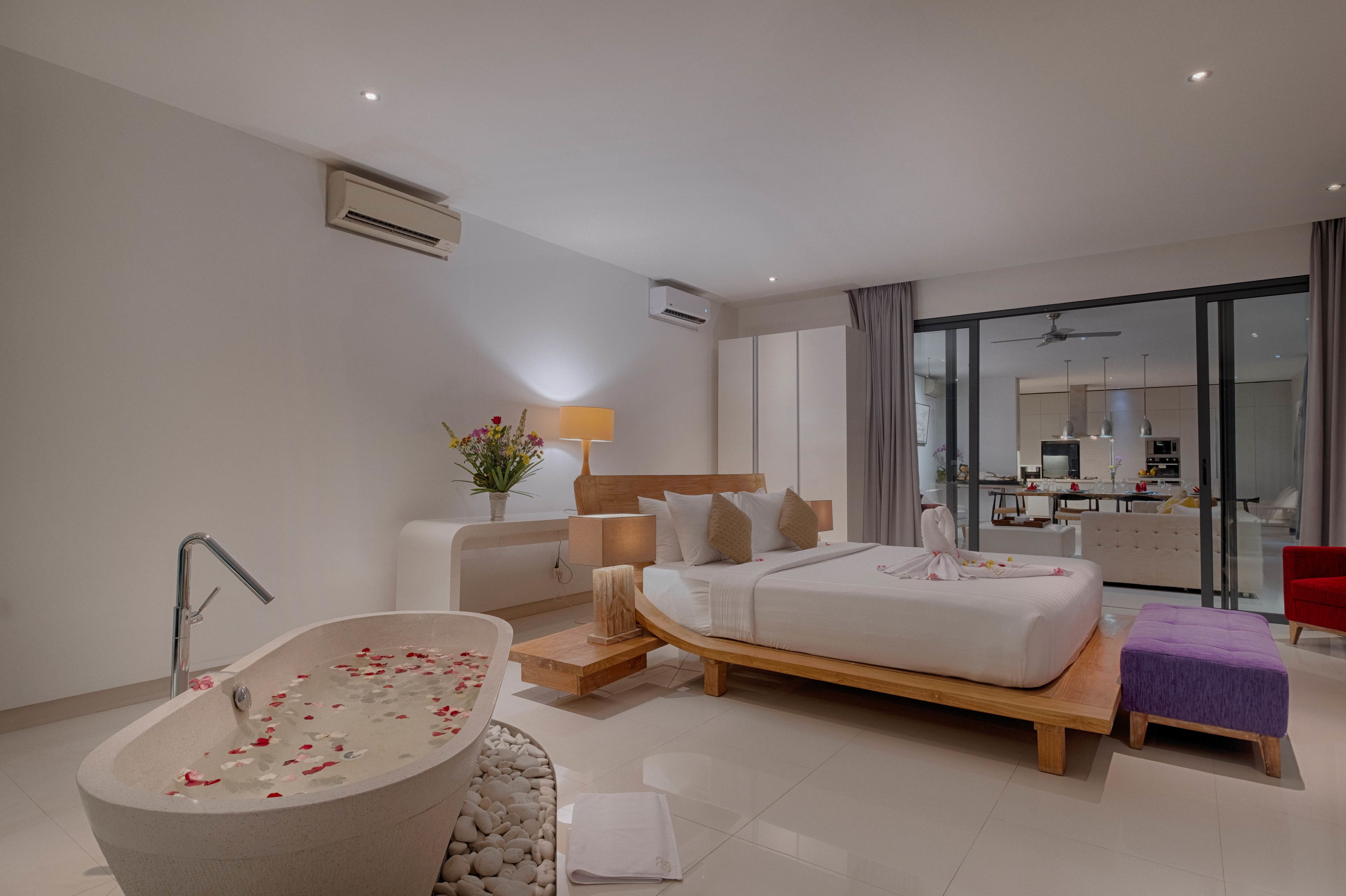 A modern hotel room with a large bathtub, a bed with a wooden frame, a small seating area, and a view of the kitchen and dining area through a glass door.