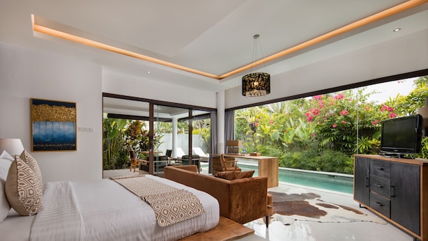 A modern bedroom with a large bed, a seating area, a TV, and a view of a pool and garden. - Canggu Villa by Sanga Sanga Hospitality (Canggu)