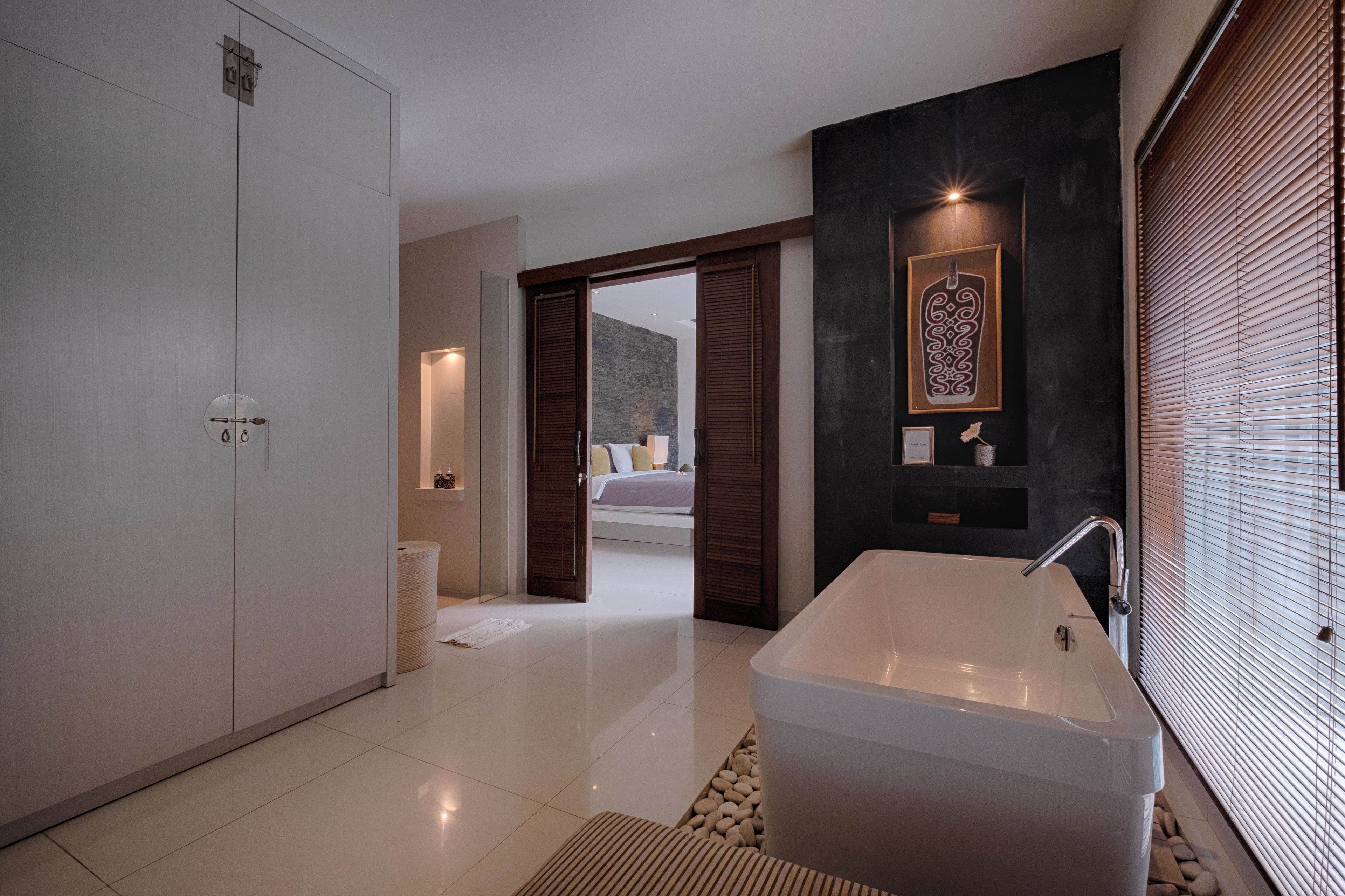 A modern bathroom with a large bathtub, a walk-in shower, and a visible bedroom with a bed and nightstand.