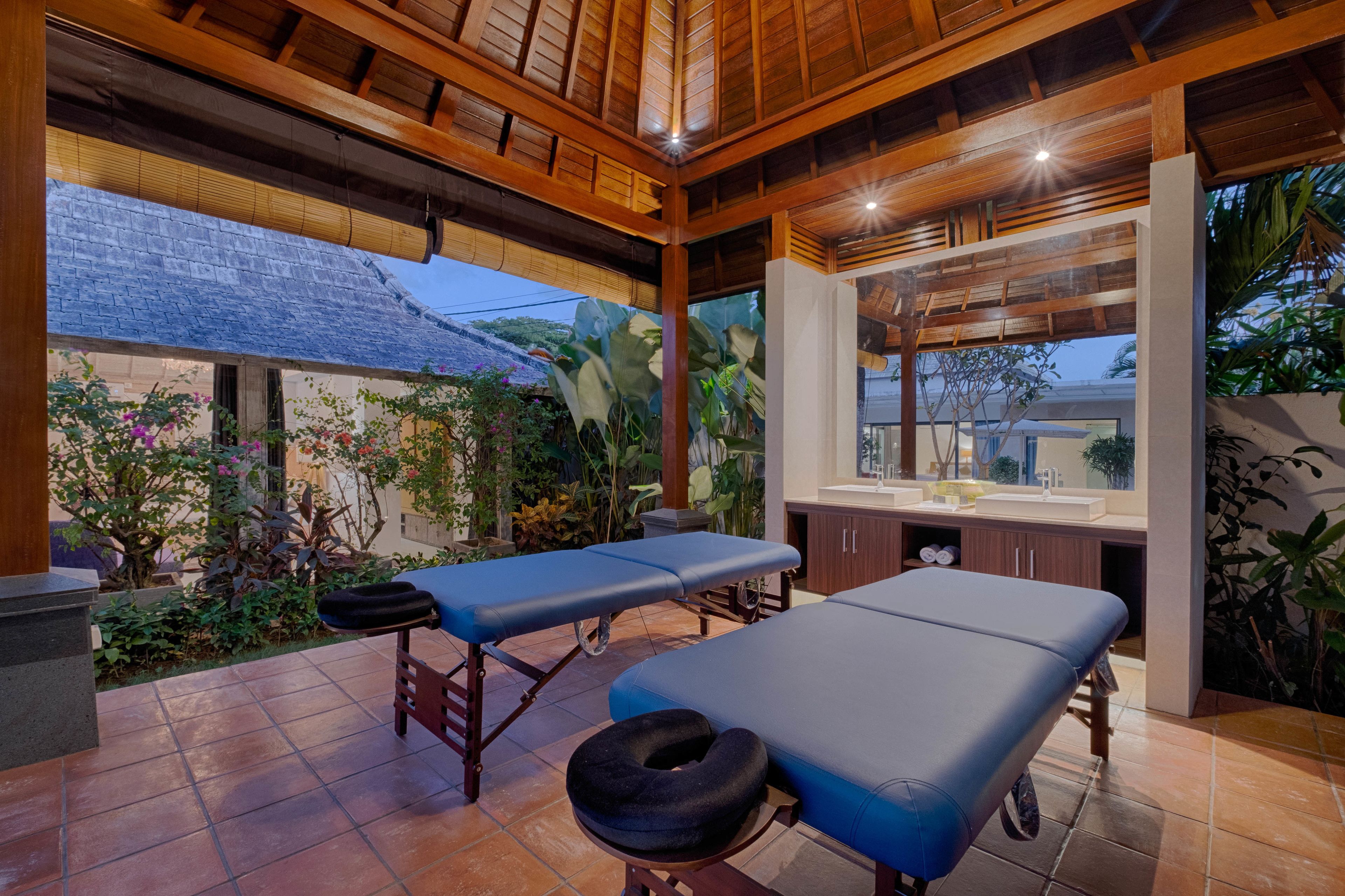 Un área de spa con dos camillas de masaje, techo de madera y vistas a un jardín con plantas y un edificio al fondo.